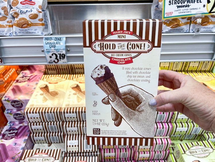 A person's hand holding a box fo Trader Joe's Mini Hold the Cone Ice Cream Cones in front of a freezer shelf inside Trader Joe's.