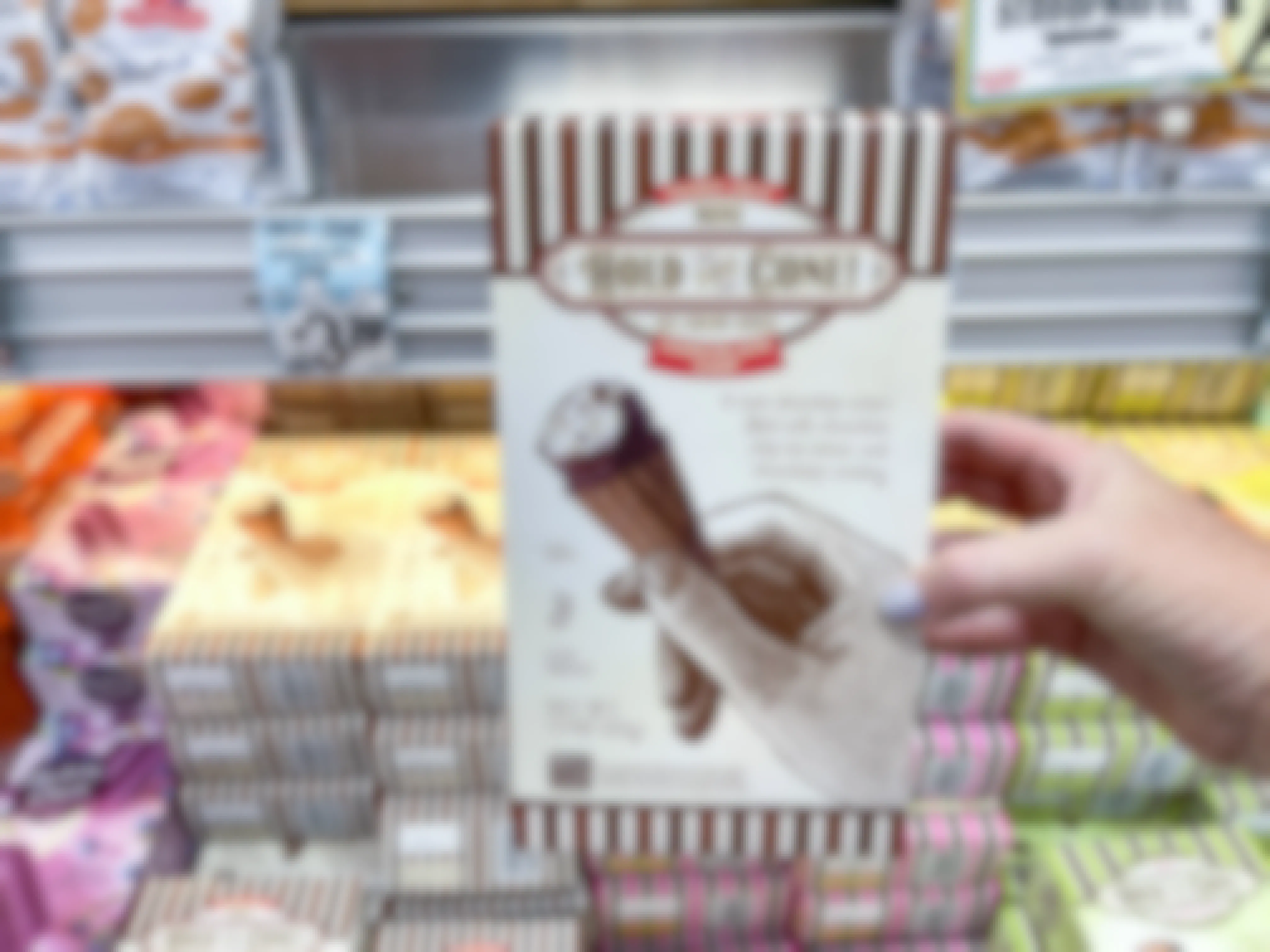 A person's hand holding a box fo Trader Joe's Mini Hold the Cone Ice Cream Cones in front of a freezer shelf inside Trader Joe's.