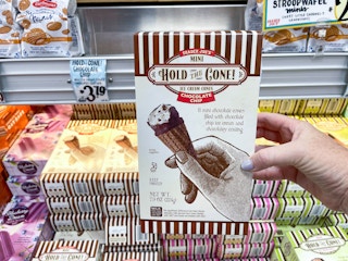 A person's hand holding a box fo Trader Joe's Mini Hold the Cone Ice Cream Cones in front of a freezer shelf inside Trader Joe's.