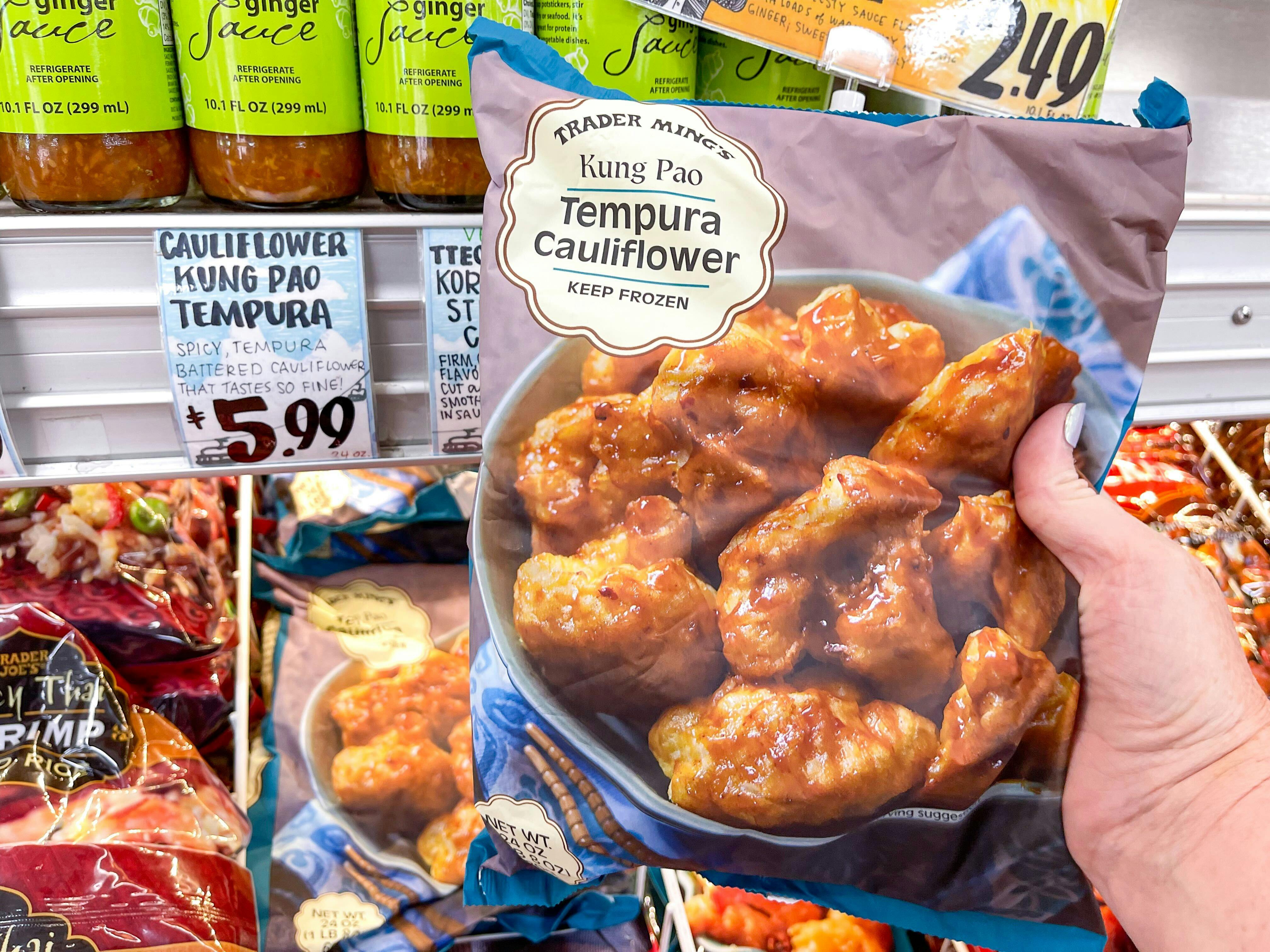 A person's hand holding a bag of Trader Ming's Kung Pao Tempura Cauliflower over a display of them in Trader Joe's.