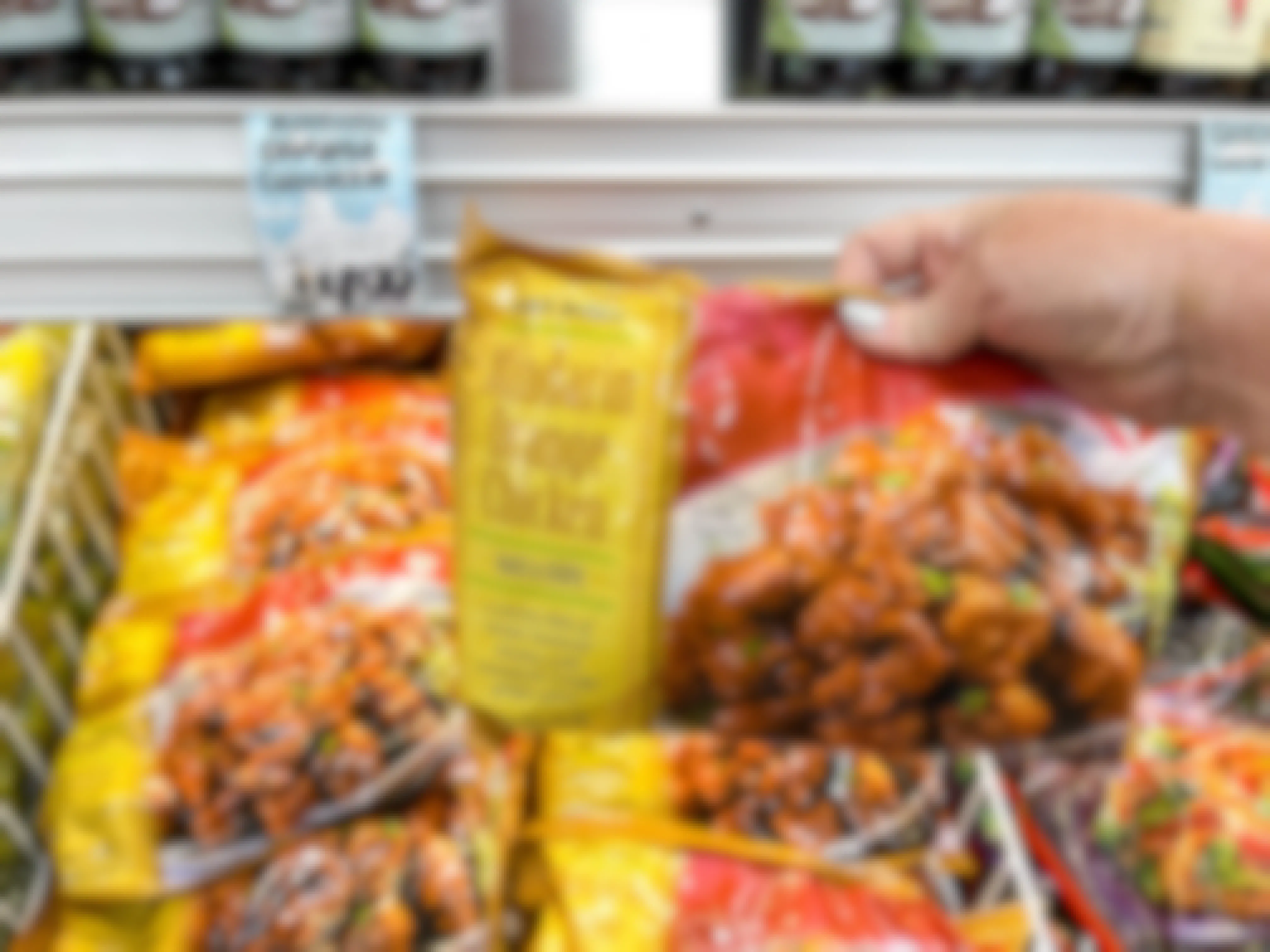 A person's hand holding a bag of Trader Ming's Mandarin Orange Chicken above a display of them at Trader Joe's.