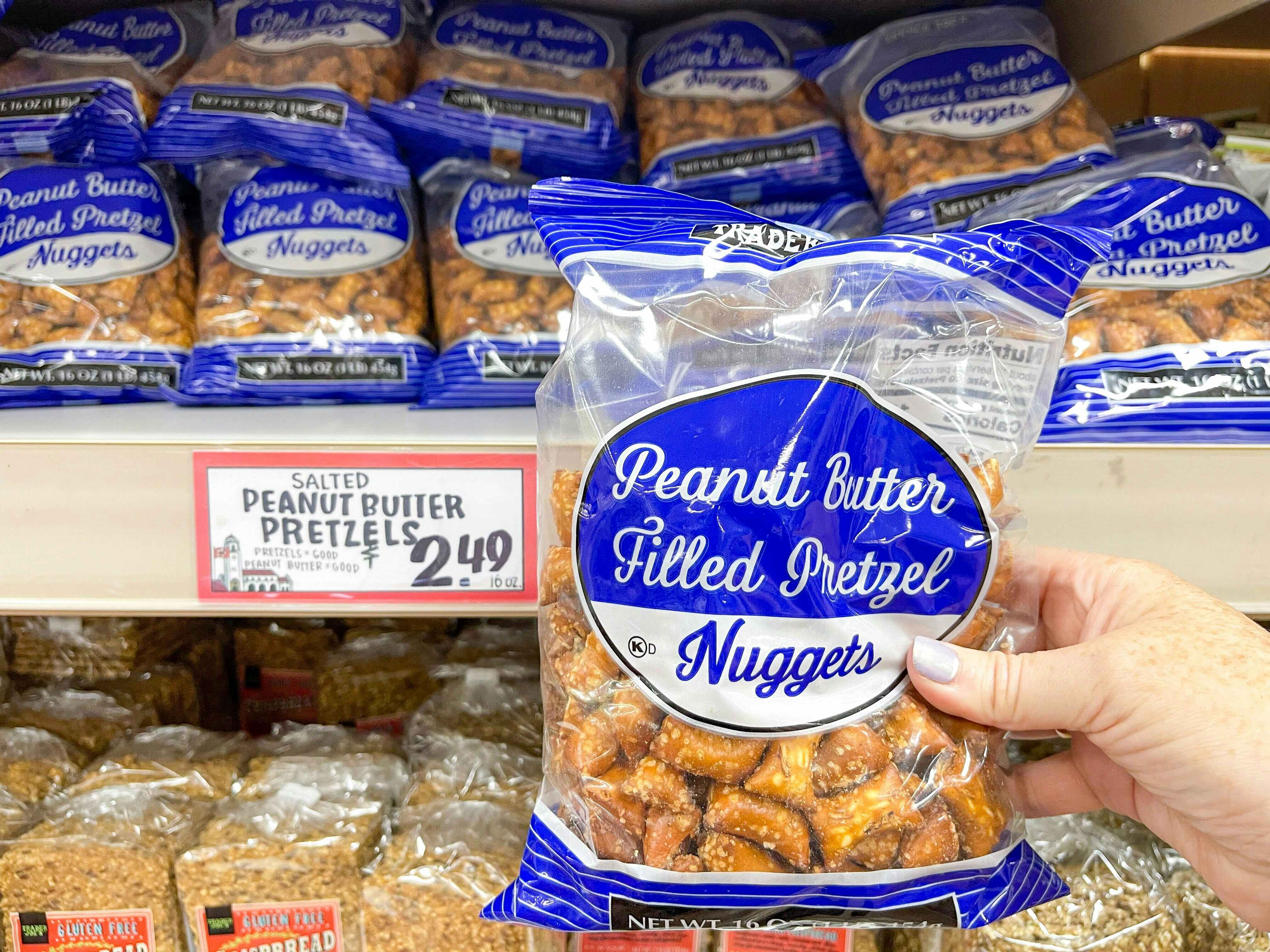 A person's hand holding a bag of Trader Joe's Peanut Butter Filled Pretzel Nuggets in front of a shelf display of them at Trader Joe's