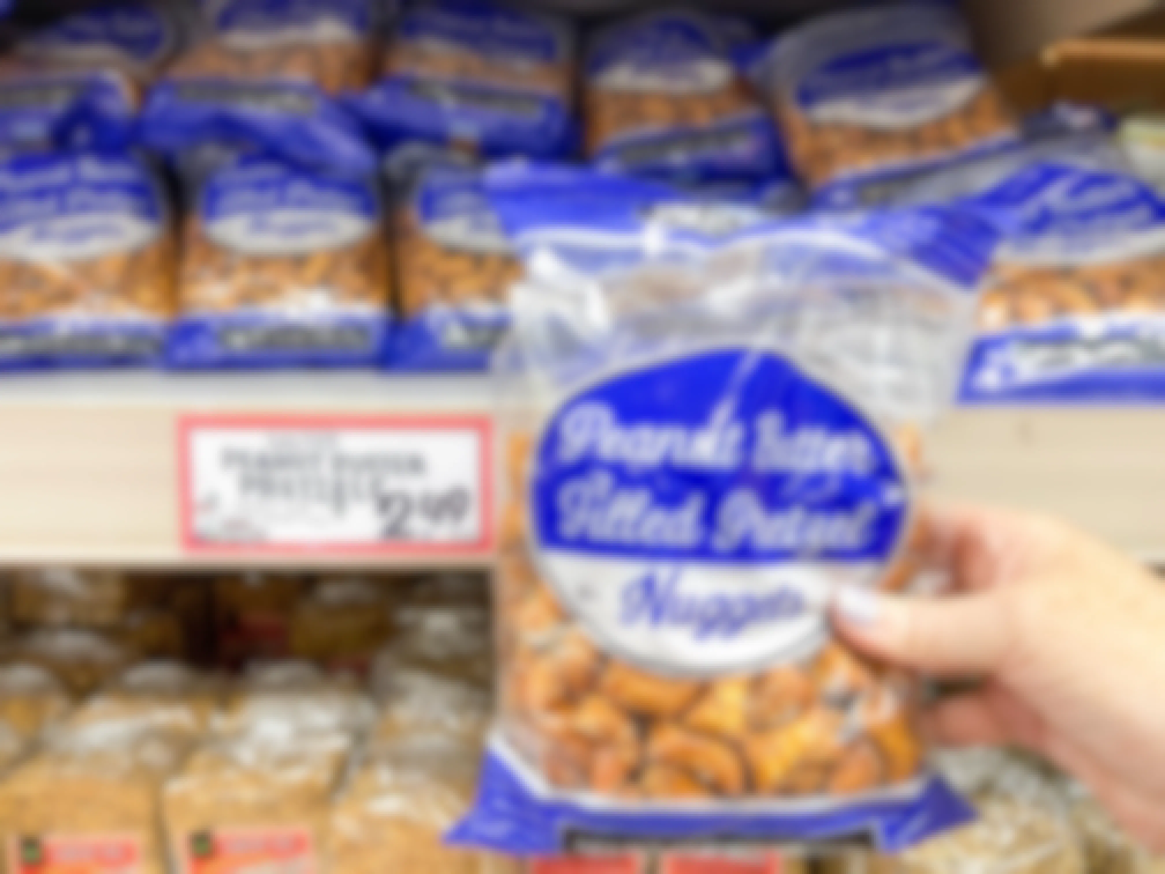 A person's hand holding a bag of Trader Joe's Peanut Butter Filled Pretzel Nuggets in front of a shelf display of them at Trader Joe's
