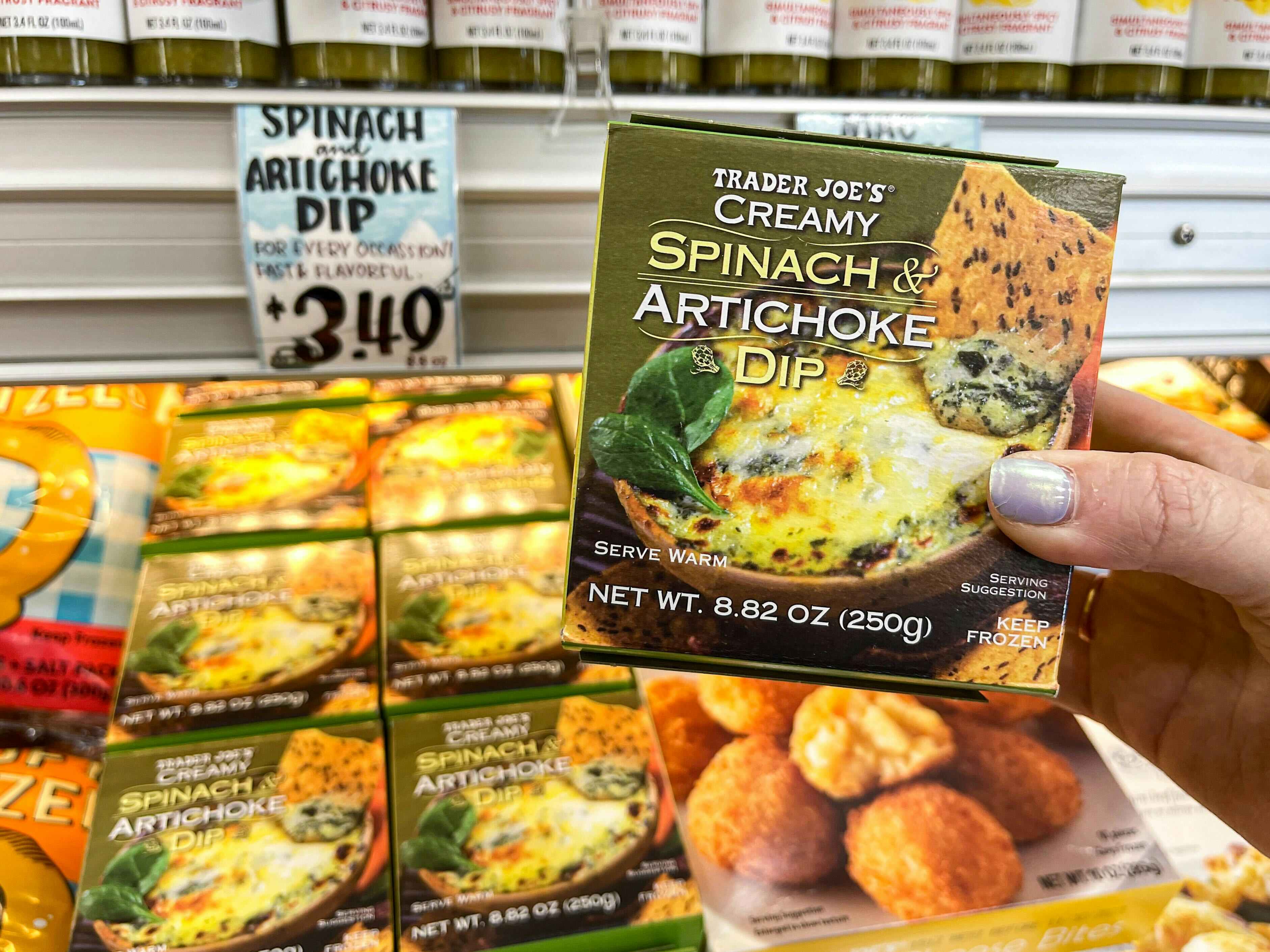 A person's hand holding a box of Trader Joe's Creamy Spinach & Artichoke Dip in front of the display and price sign at Trader Joe's.