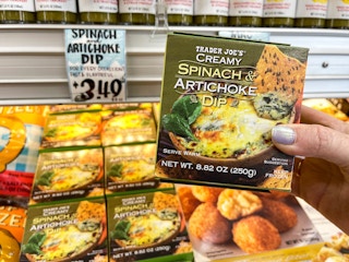 A person's hand holding a box of Trader Joe's Creamy Spinach & Artichoke Dip in front of the display and price sign at Trader Joe's.