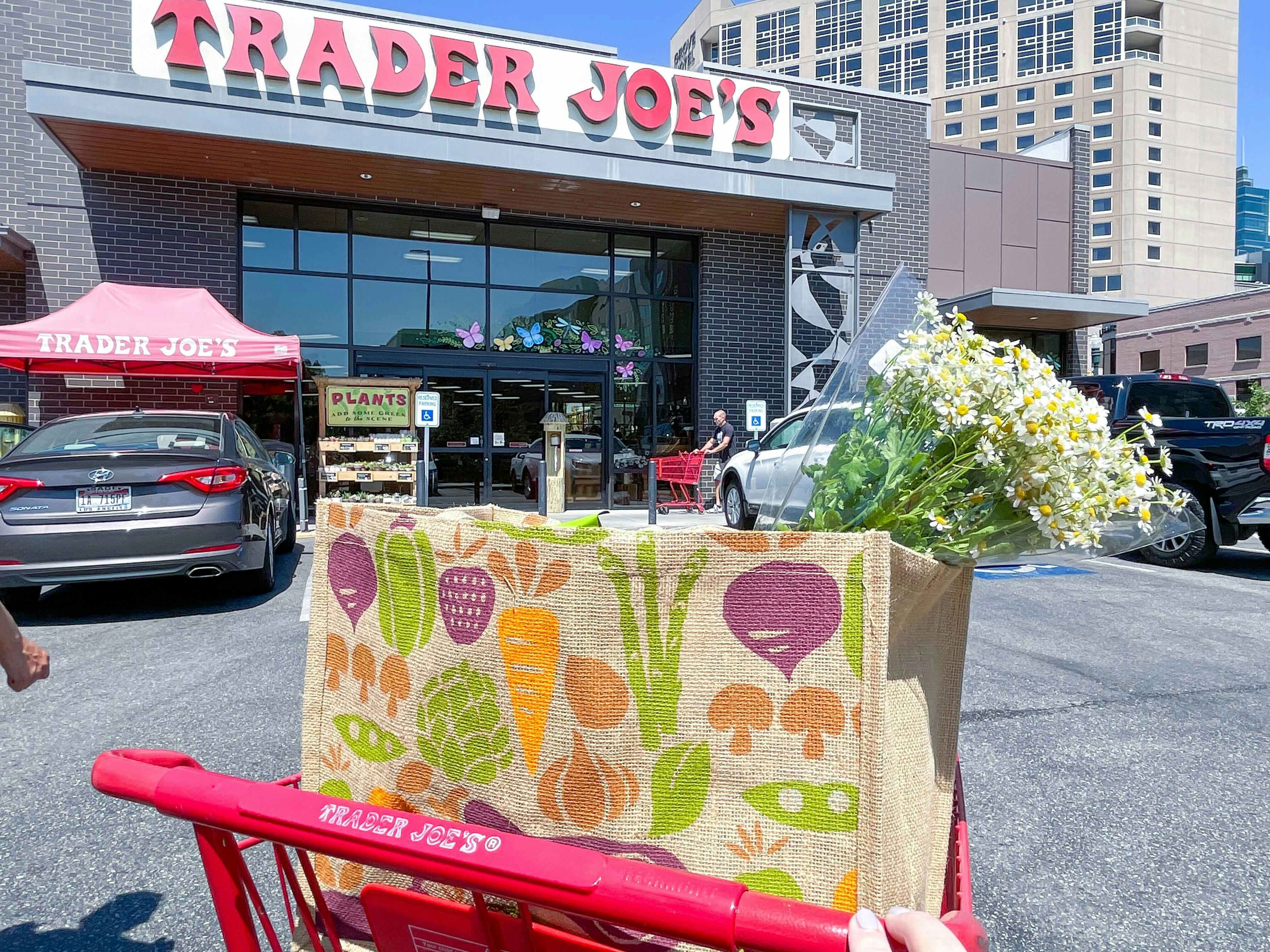 A Trader Joe's shopping cart with bags of groceries in the basket, parked outside of a Trader Joe's grocery store.
