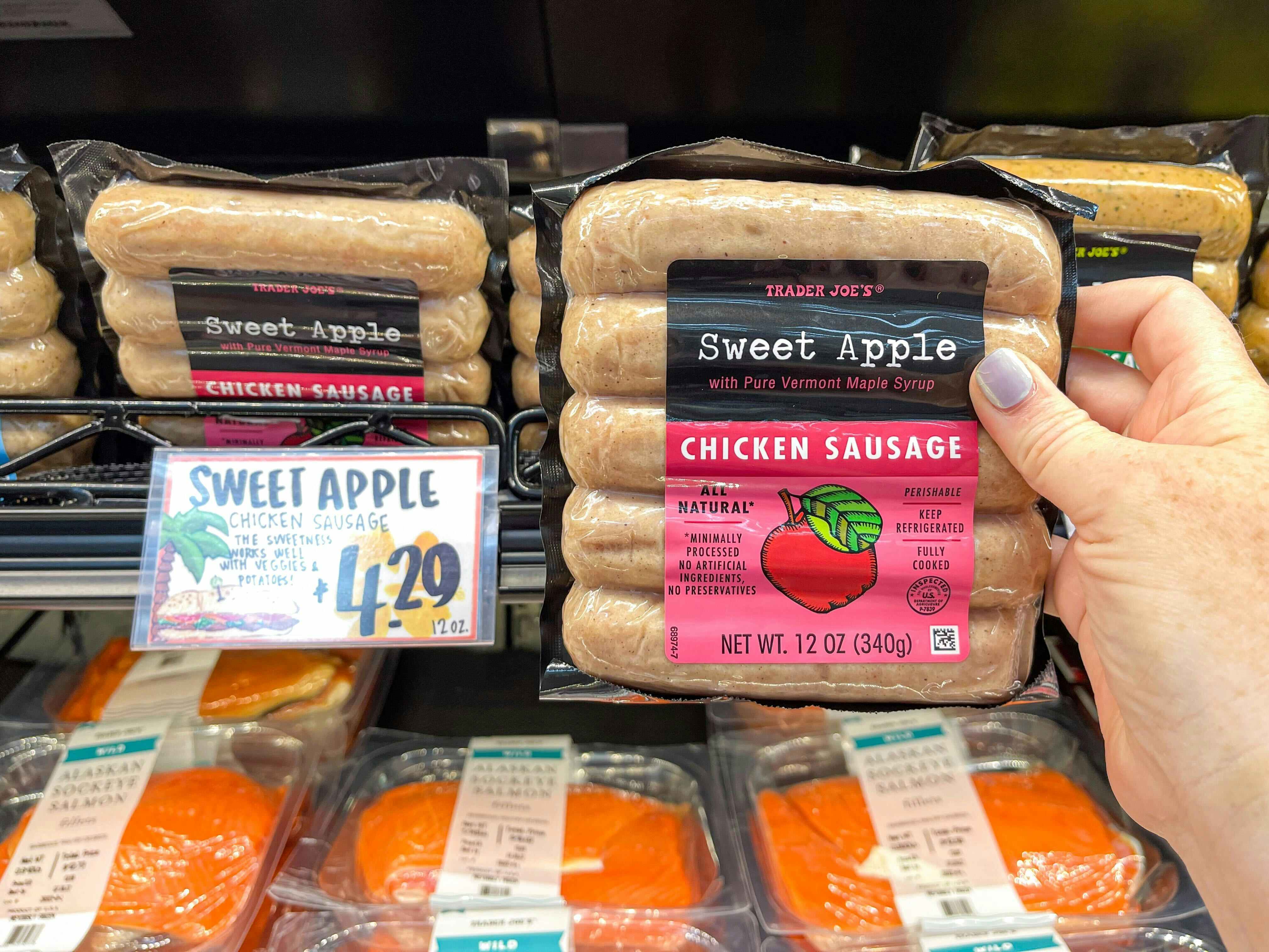 A person's hand holding a package of Trader Joe's Sweet Apple Chicken Sausage near a price sign on a refrigerated shelf at Trader Joe's.