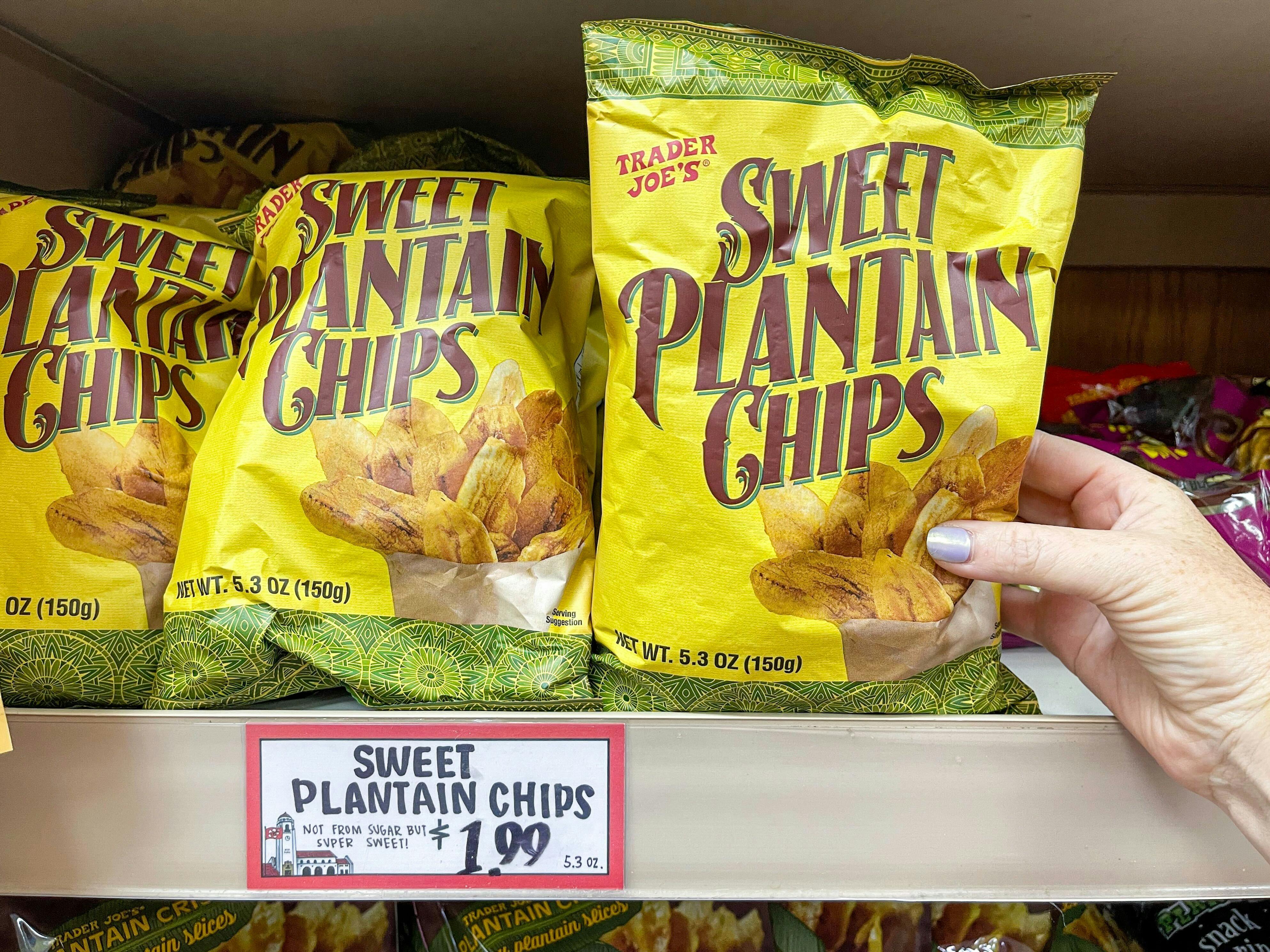 A person's hand taking a bag of Trader Joe's Sweet Plantain Chips of off a shelf of them at Trader Joe's.