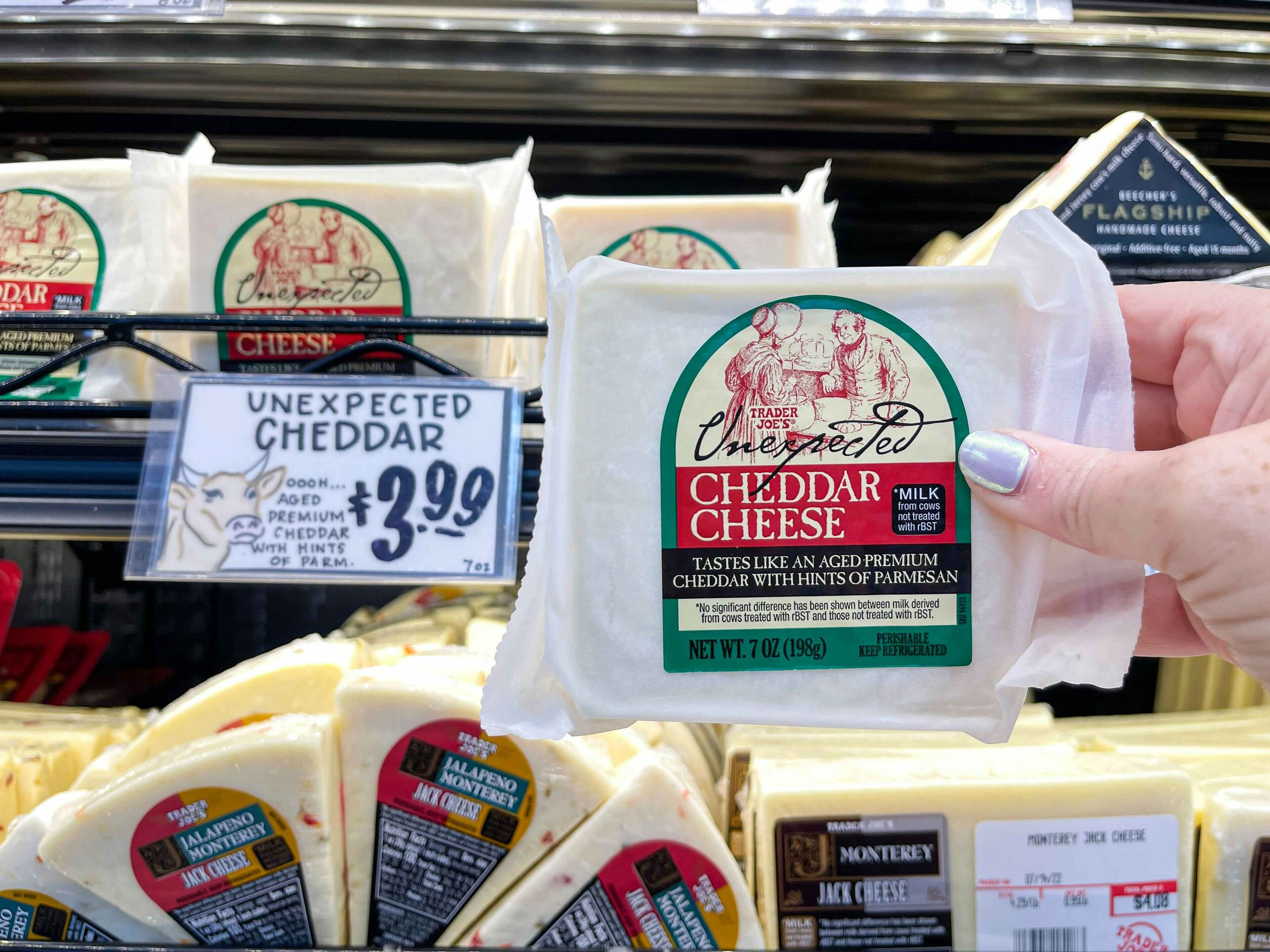 A person's hand holding a package of Trader Joe's Unexpected Cheddar Cheese in front of the price sign on the refrigerated shelf at Trader Joe's..
