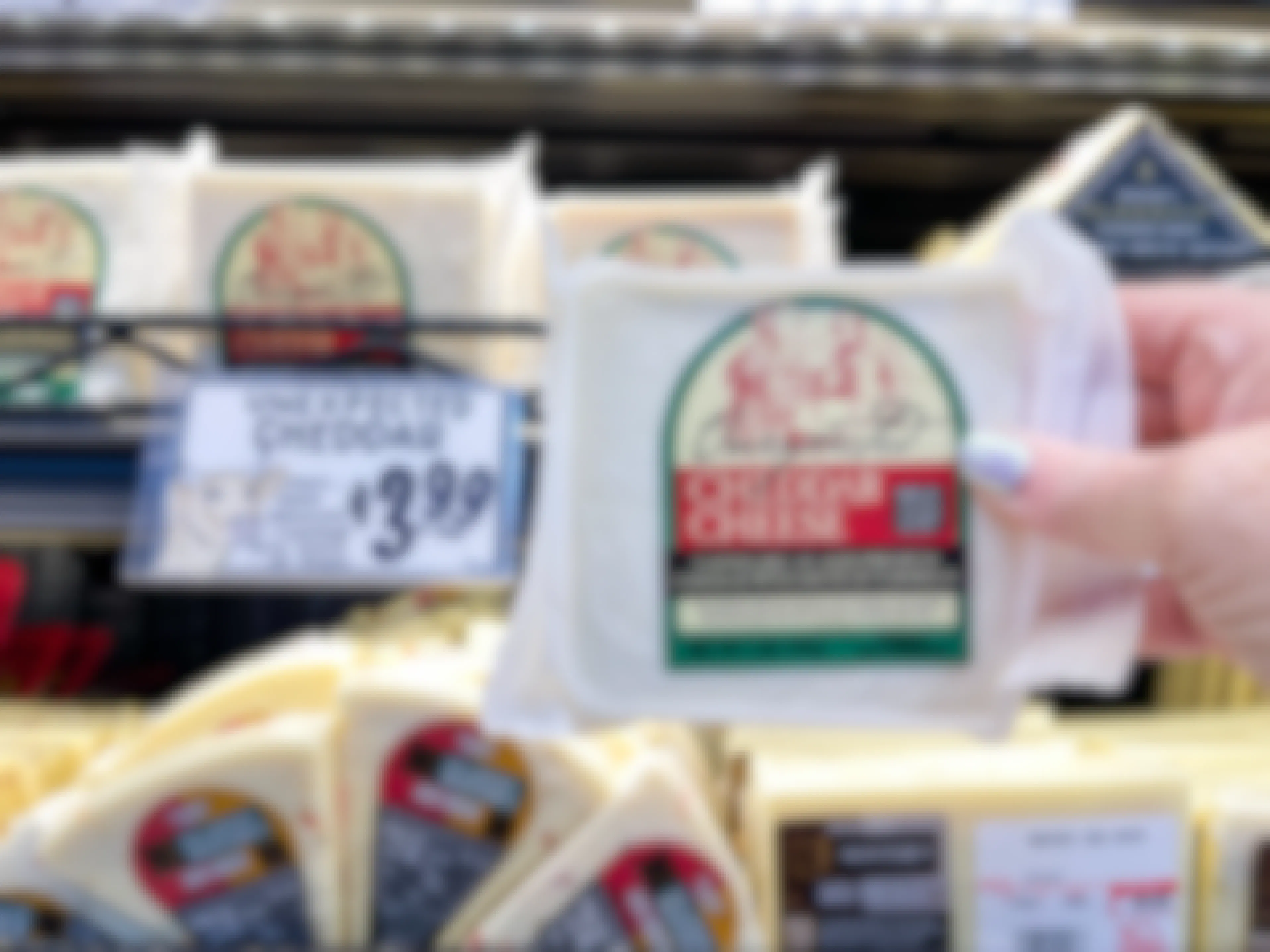 A person's hand holding a package of Trader Joe's Unexpected Cheddar Cheese in front of the price sign on the refrigerated shelf at Trader Joe's..