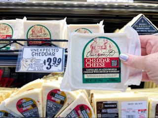 A person's hand holding a package of Trader Joe's Unexpected Cheddar Cheese in front of the price sign on the refrigerated shelf at Trader Joe's..