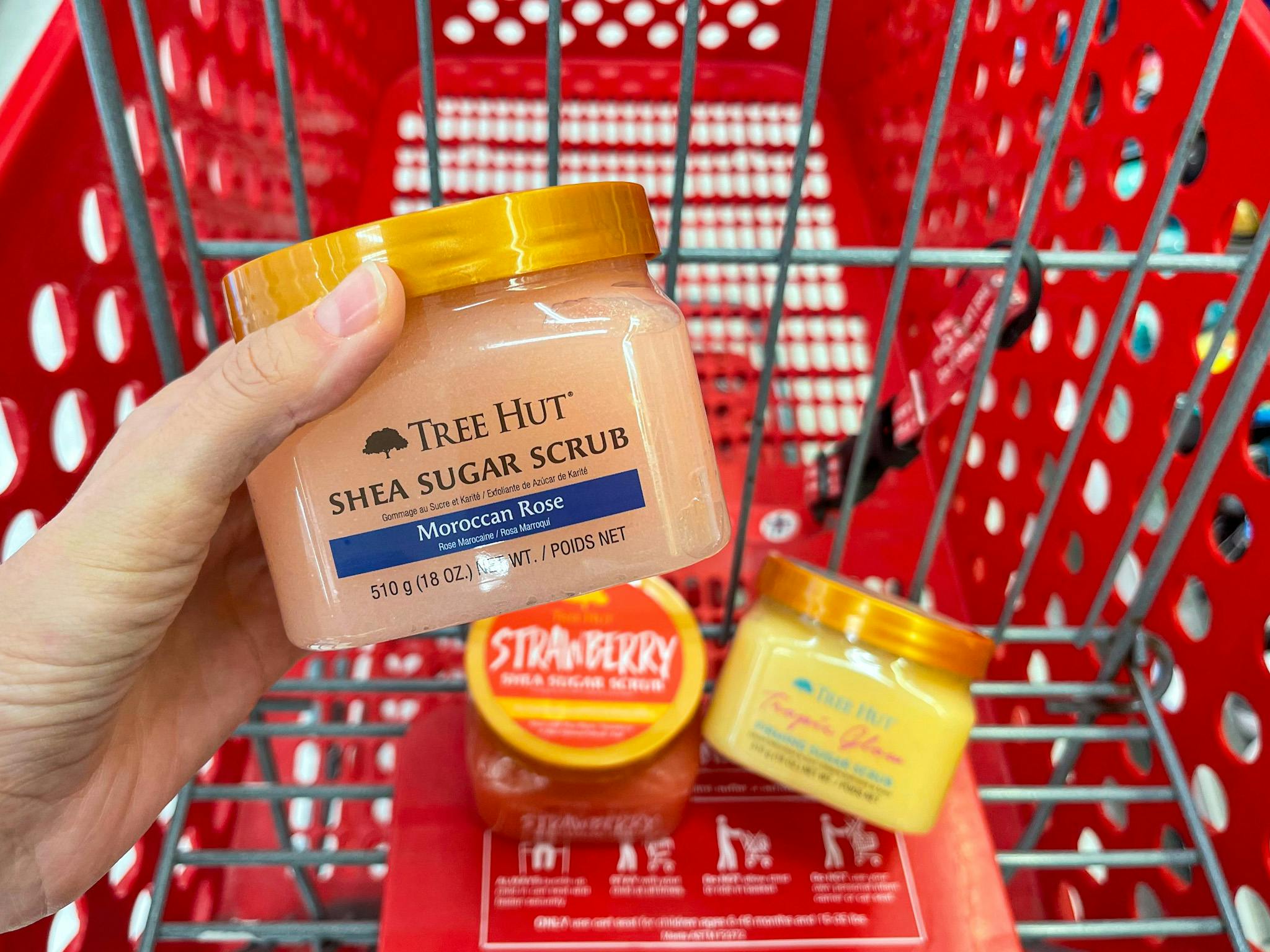 A tree hut shea sugar scrub held with hand in front of a Target shopping cart with two other jars of tree hut shea sugar scrub in it.