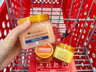 A tree hut shea sugar scrub held with hand in front of a Target shopping cart with two other jars of tree hut shea sugar scrub in it.