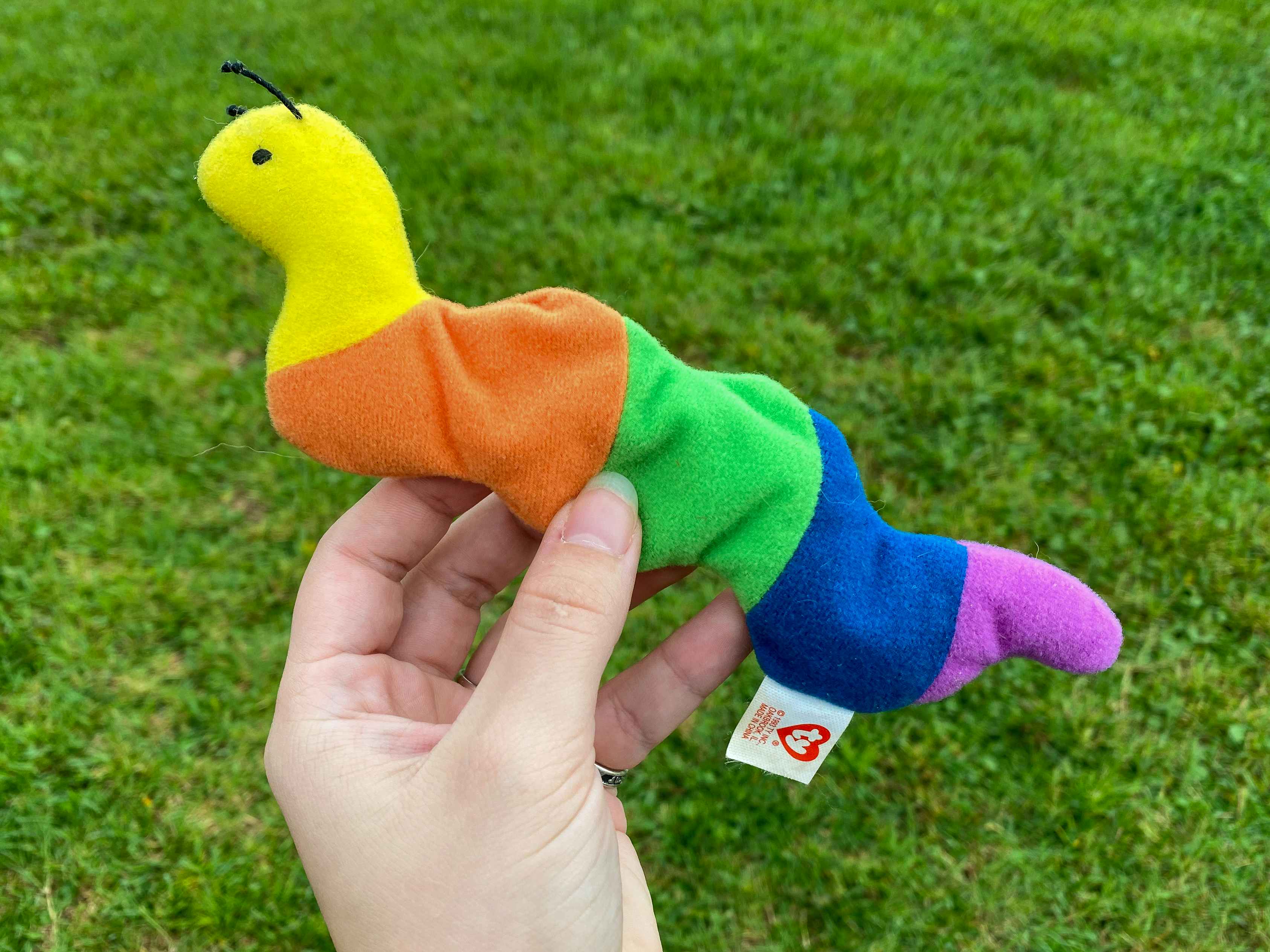A person's hand holding up an Inch the Inchworm beanie baby outside with grass in the background.