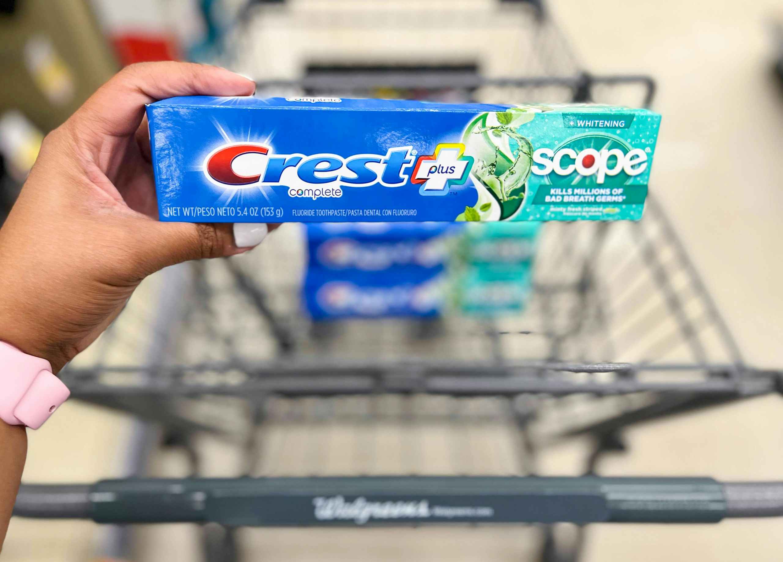 hand holding a box of Crest plus Scope toothpaste in front of shopping cart with two more boxes inside