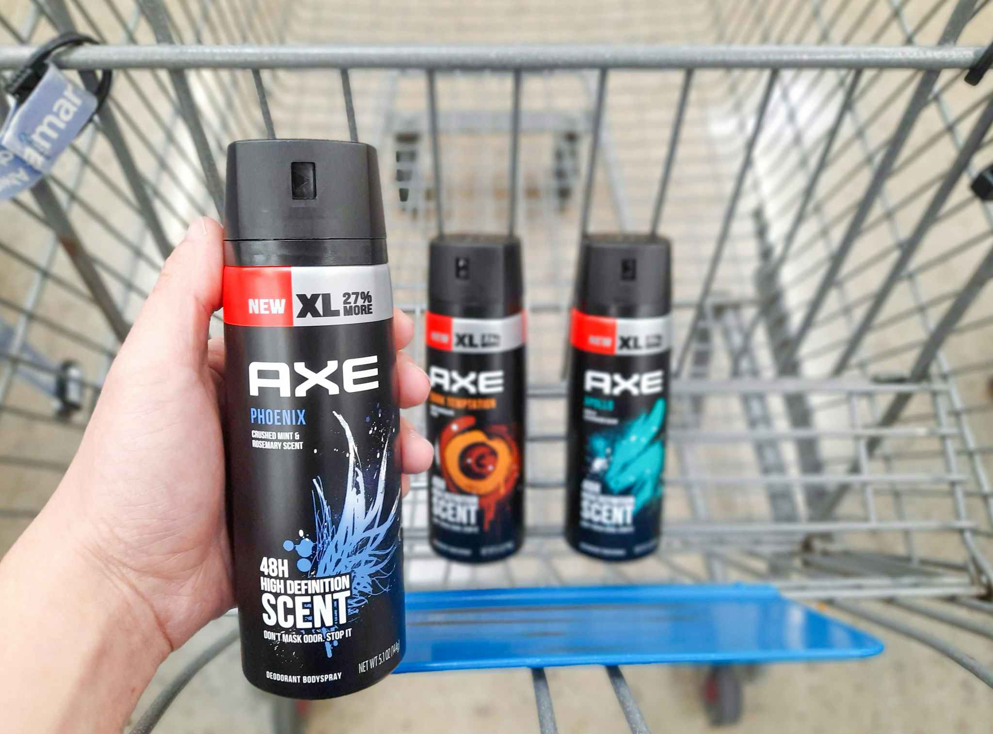 Hand holding one Axe Deodorant Body Spray product over Walmart shopping cart. Shopping cart has two Axe Deodorant Body Spray products in it.