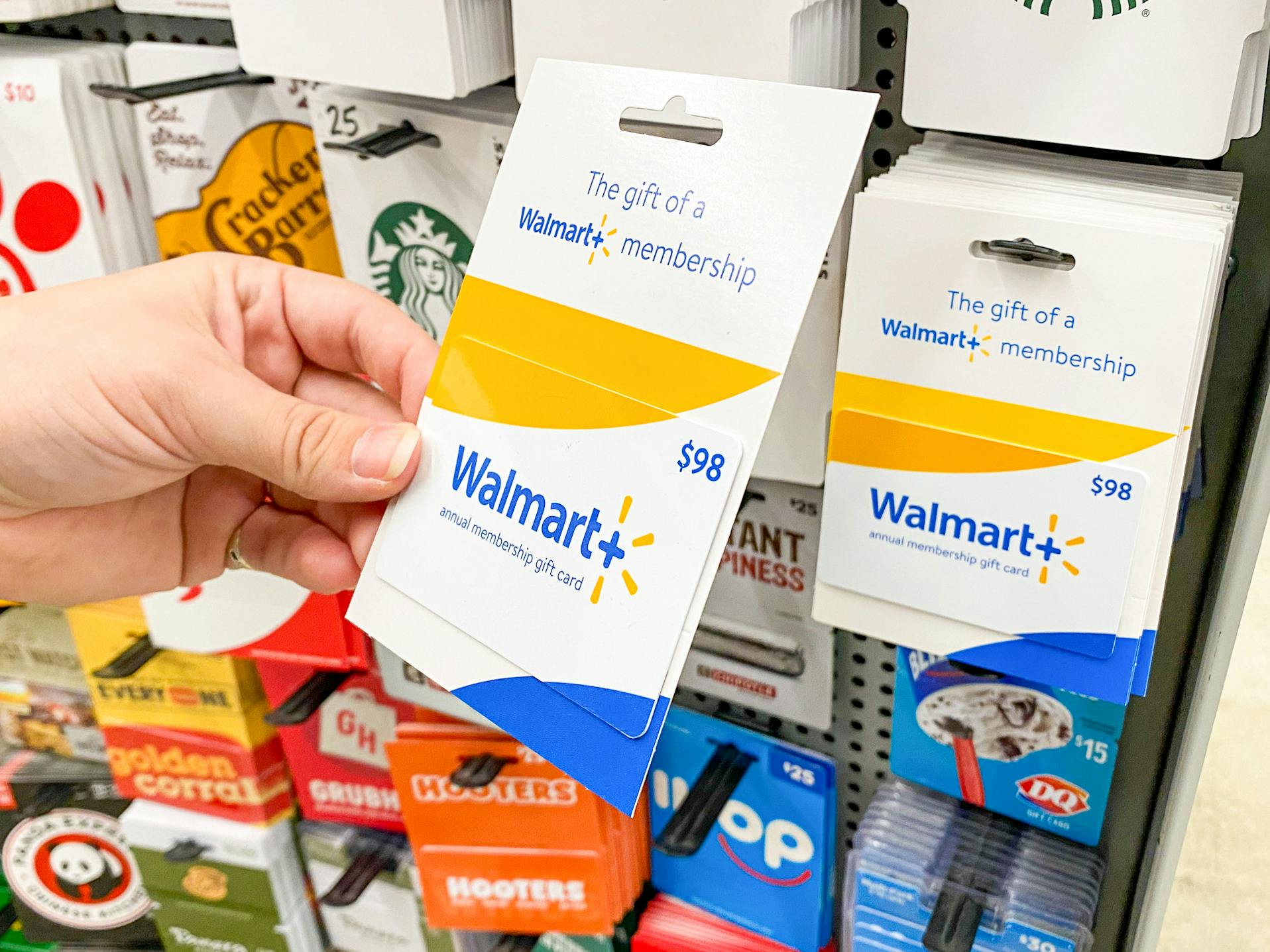 A person's hand taking a Walmart Plus gift card from a display of gift cards at Walmart.