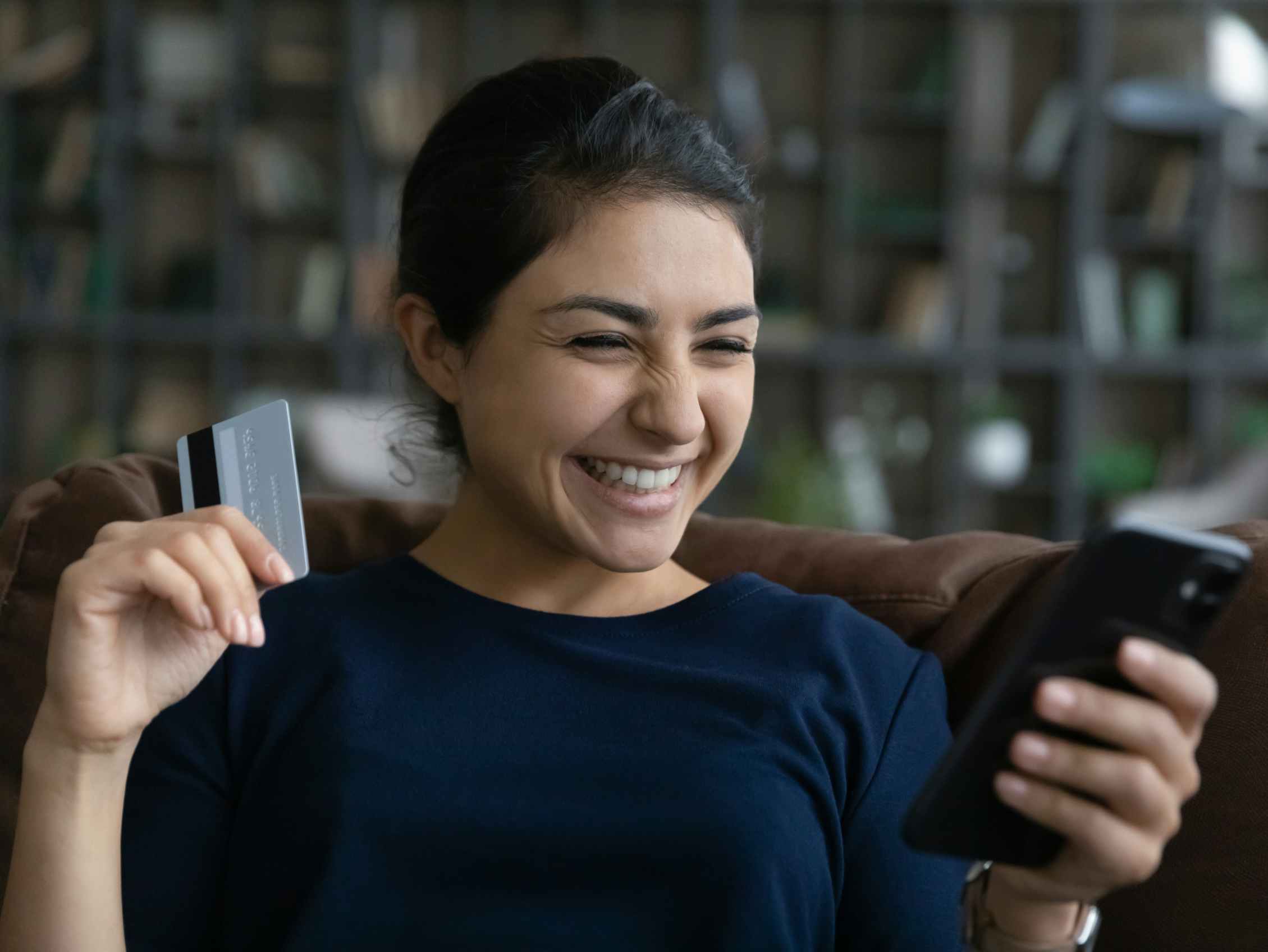 A woman grinning at her phone in one hand while holding a credit card in the other hand.