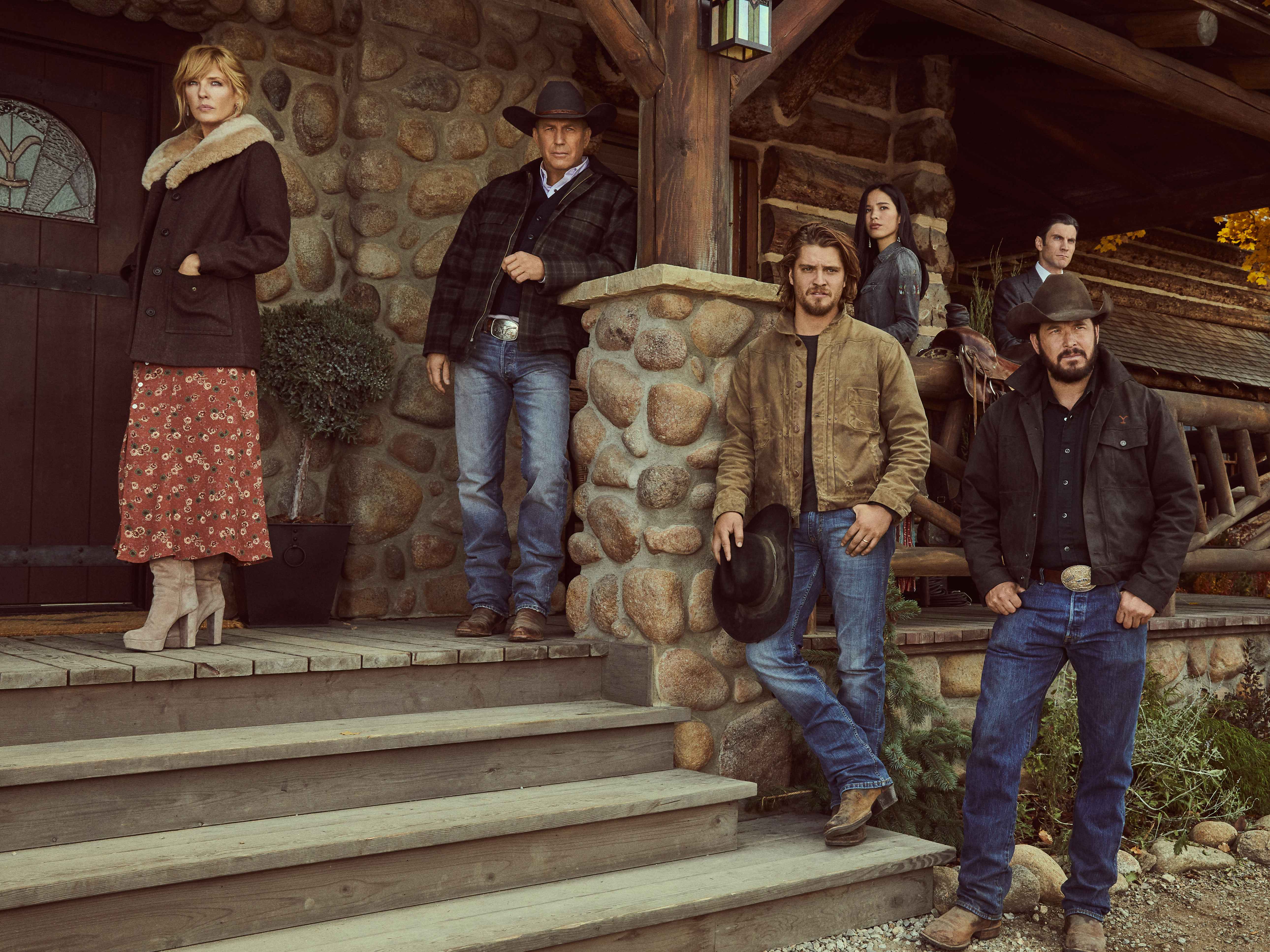 Cast members of Yellowstone posed around a front porch of a farmhouse.