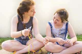 Two young girls sitting outside and working on making friendship bracelets with embroidery floss.