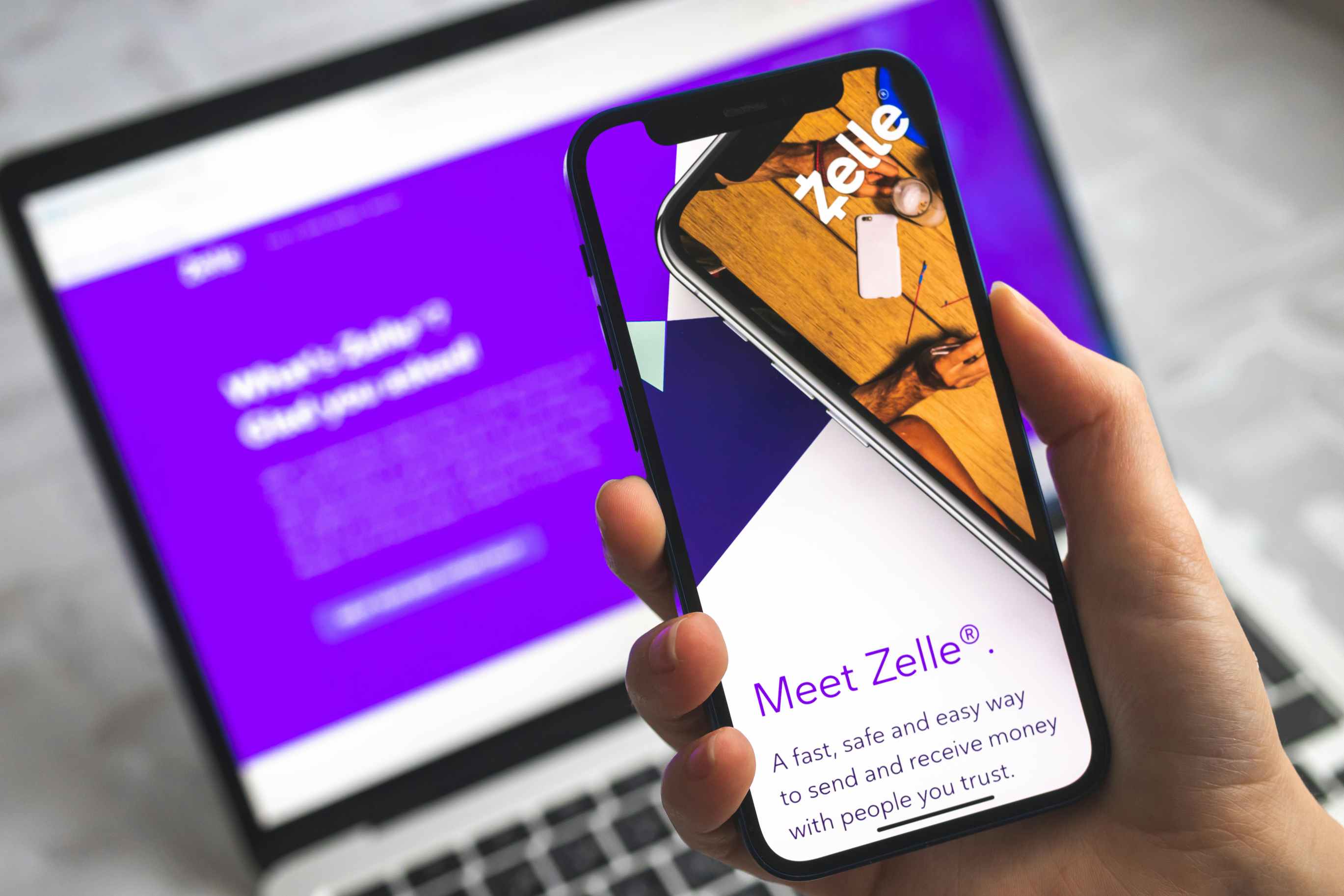 Person holding a cell phone with the Zelle payment app displayed, in front of a laptop with Zelle on it.