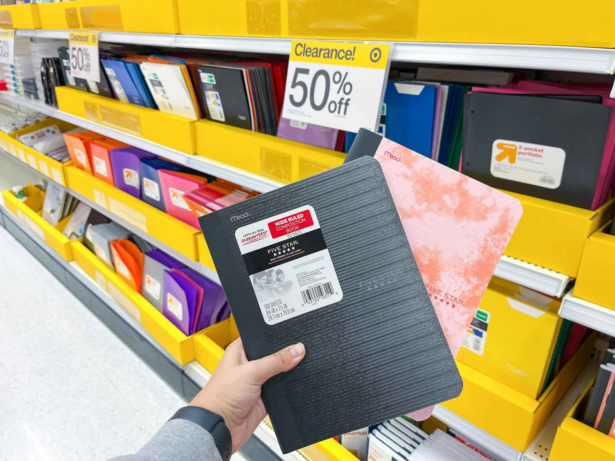 a woman's hand holding two school notebooks in front of a 50% off clearance sign on various notebooks and binders