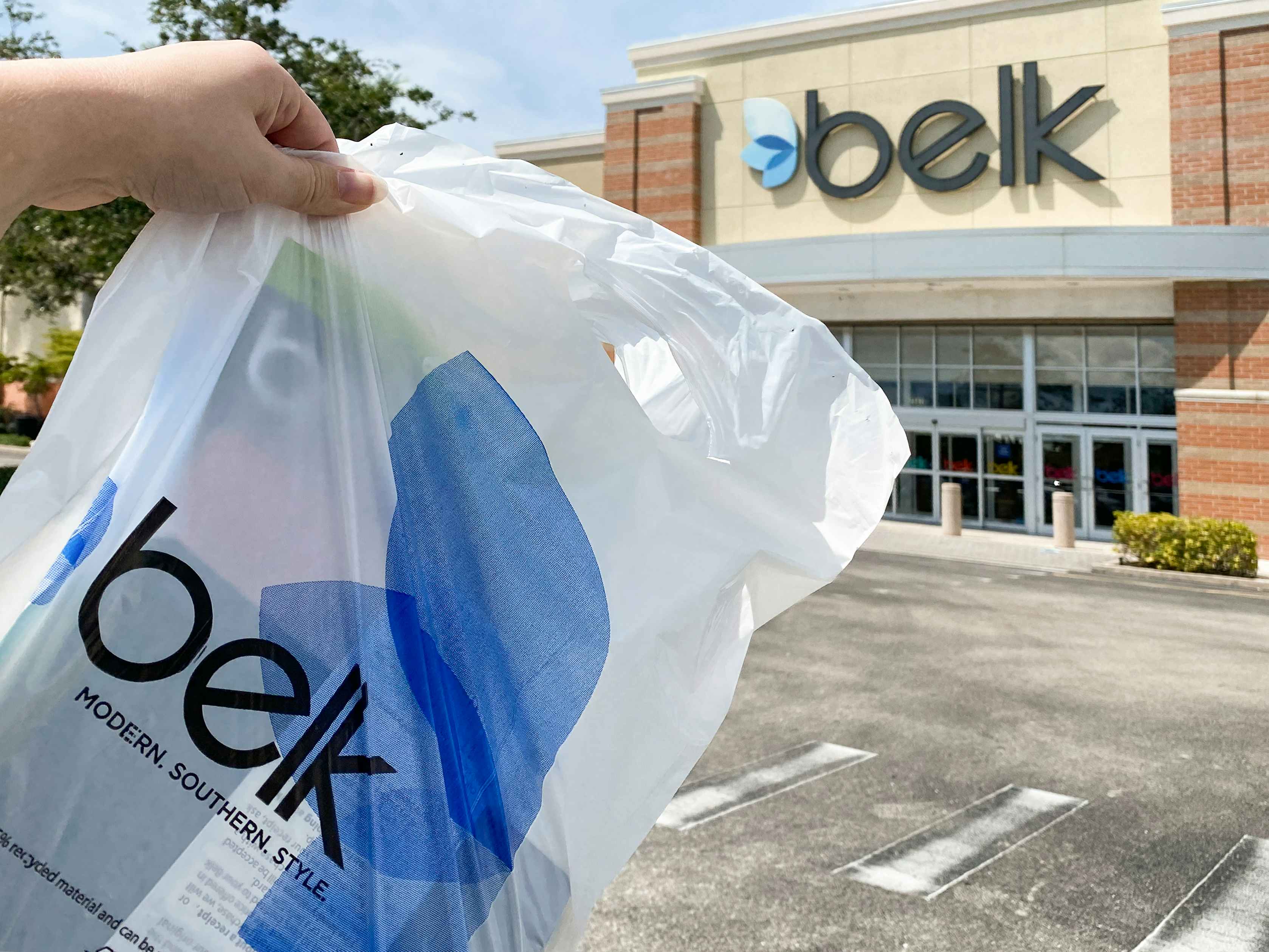 A person's hand holding up a Belk bag in front of a Belk store.