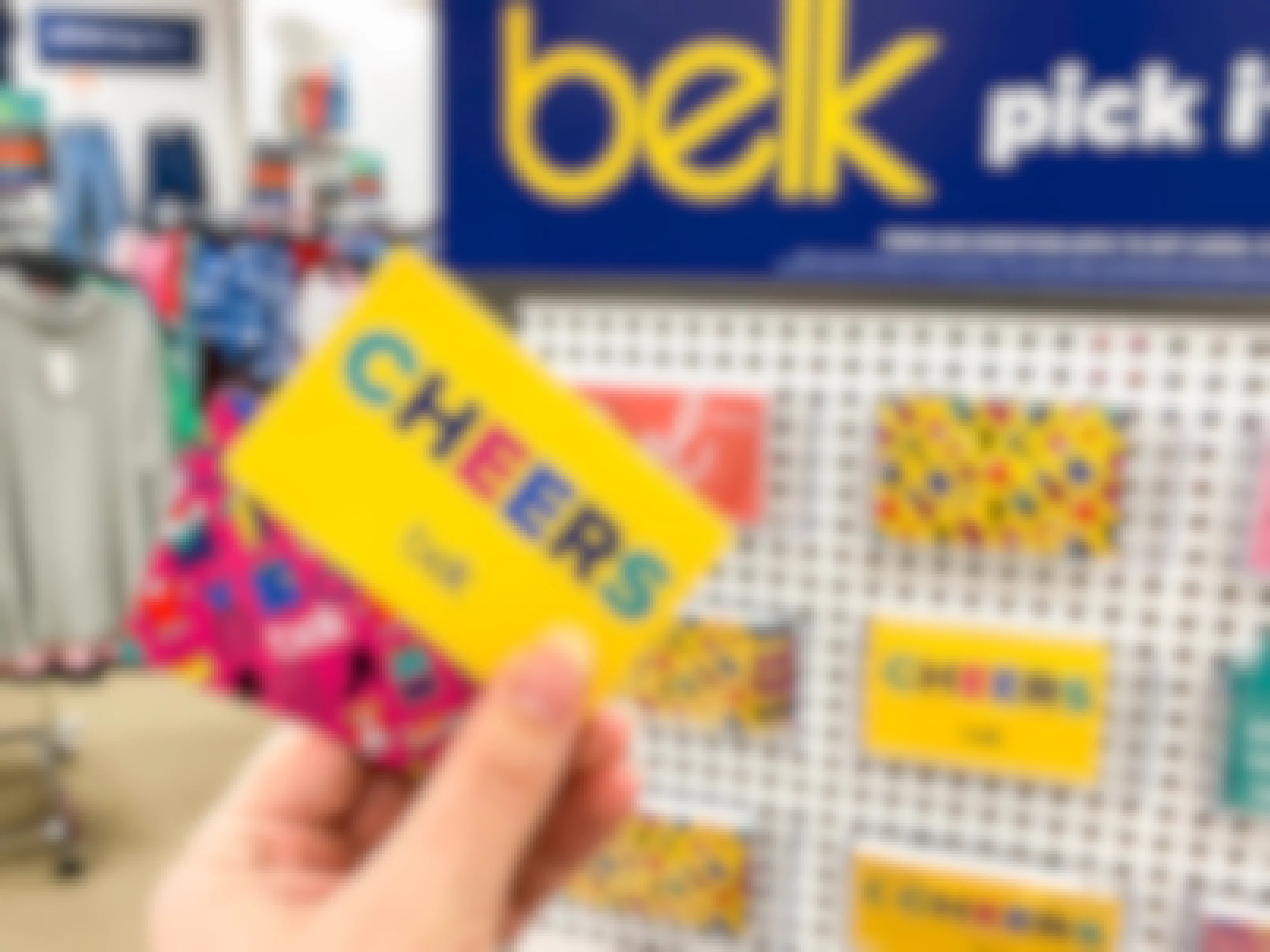 A person's hand holding some Belk gift cards in front of a display of gift cards inside Belk.