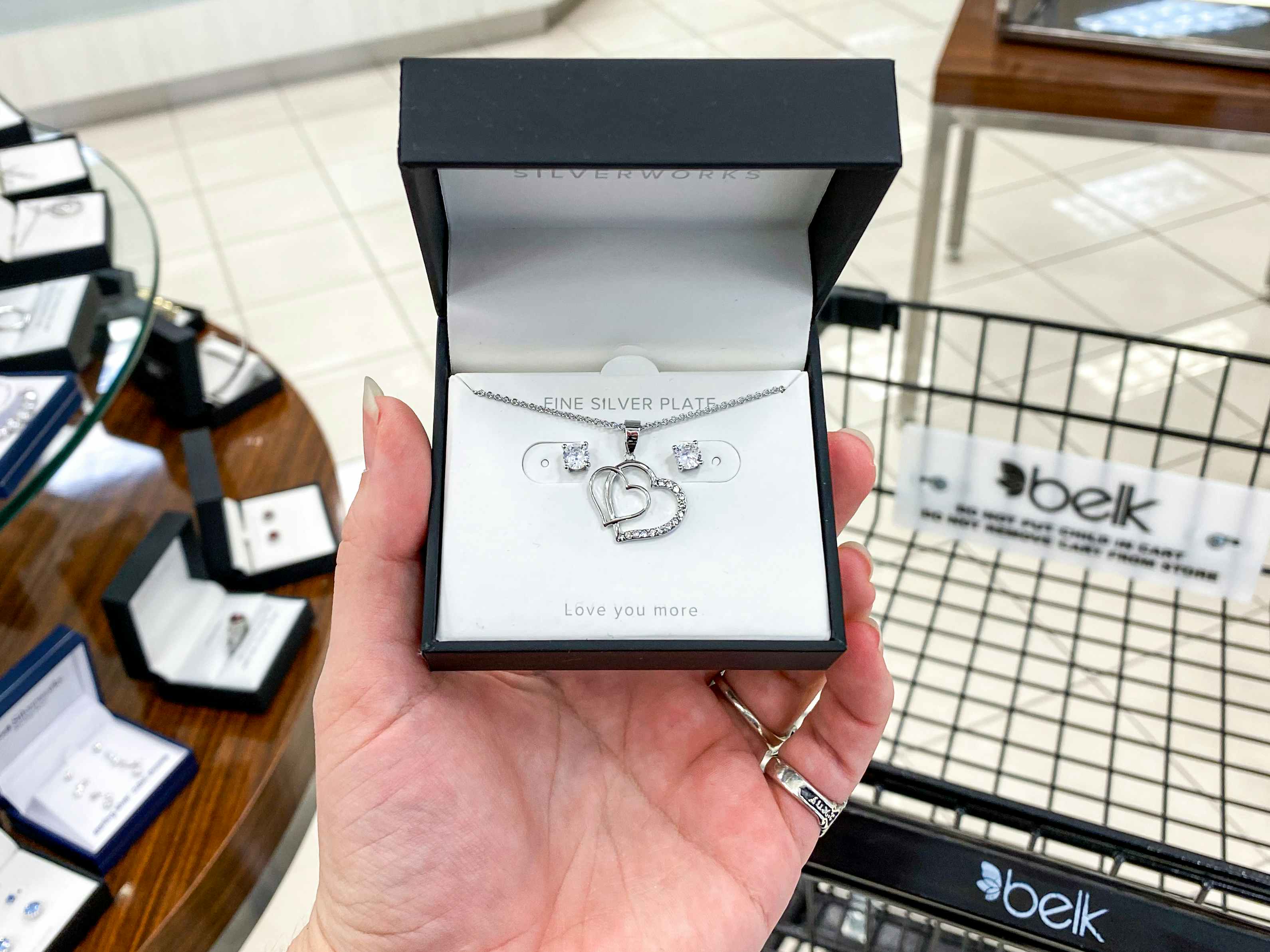 A person's hand holding a jewelry set box in front of a shelf and cart at Belk.