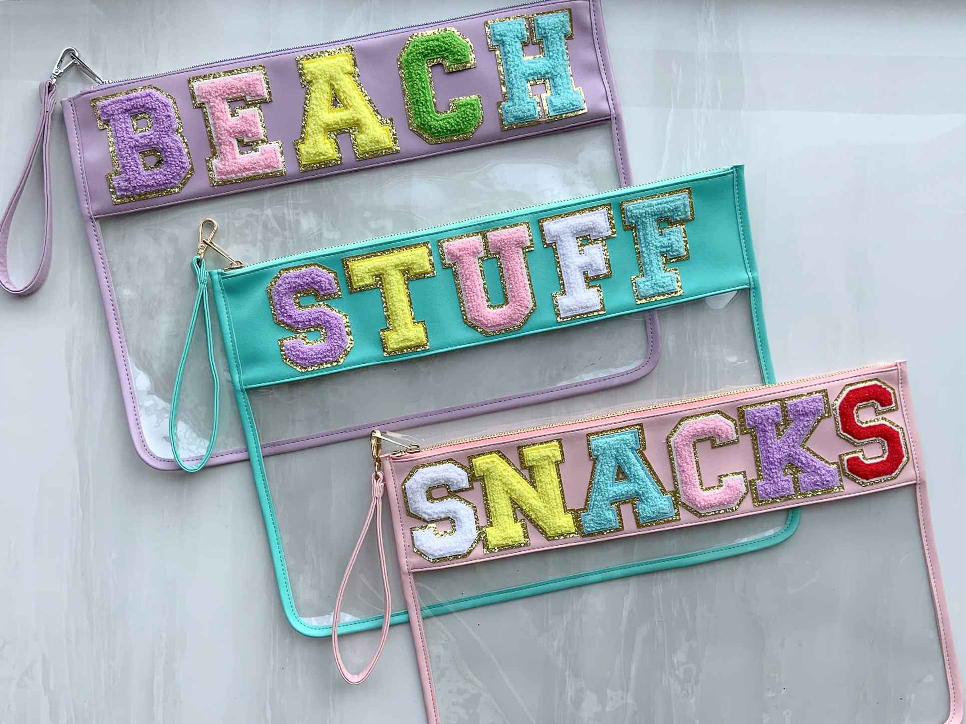 Three clear pouches customized with letter patches to say "Beach", "Stuff", and "Snacks" lying together on a marble counter.