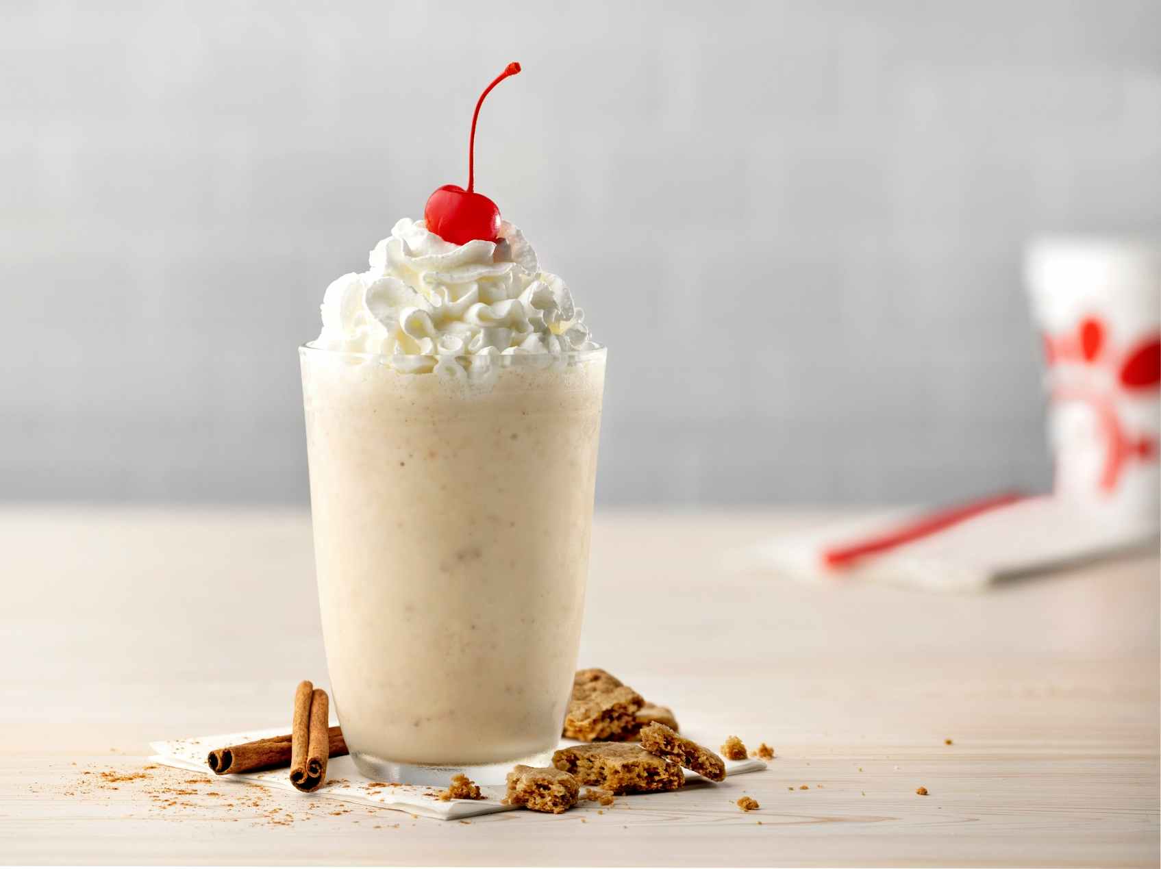 A Chick-fil-A Autumn Spice Milkshake with whipped cream and a cherry on top with cinnamon sticks and cookie crumbles on the table next to it.