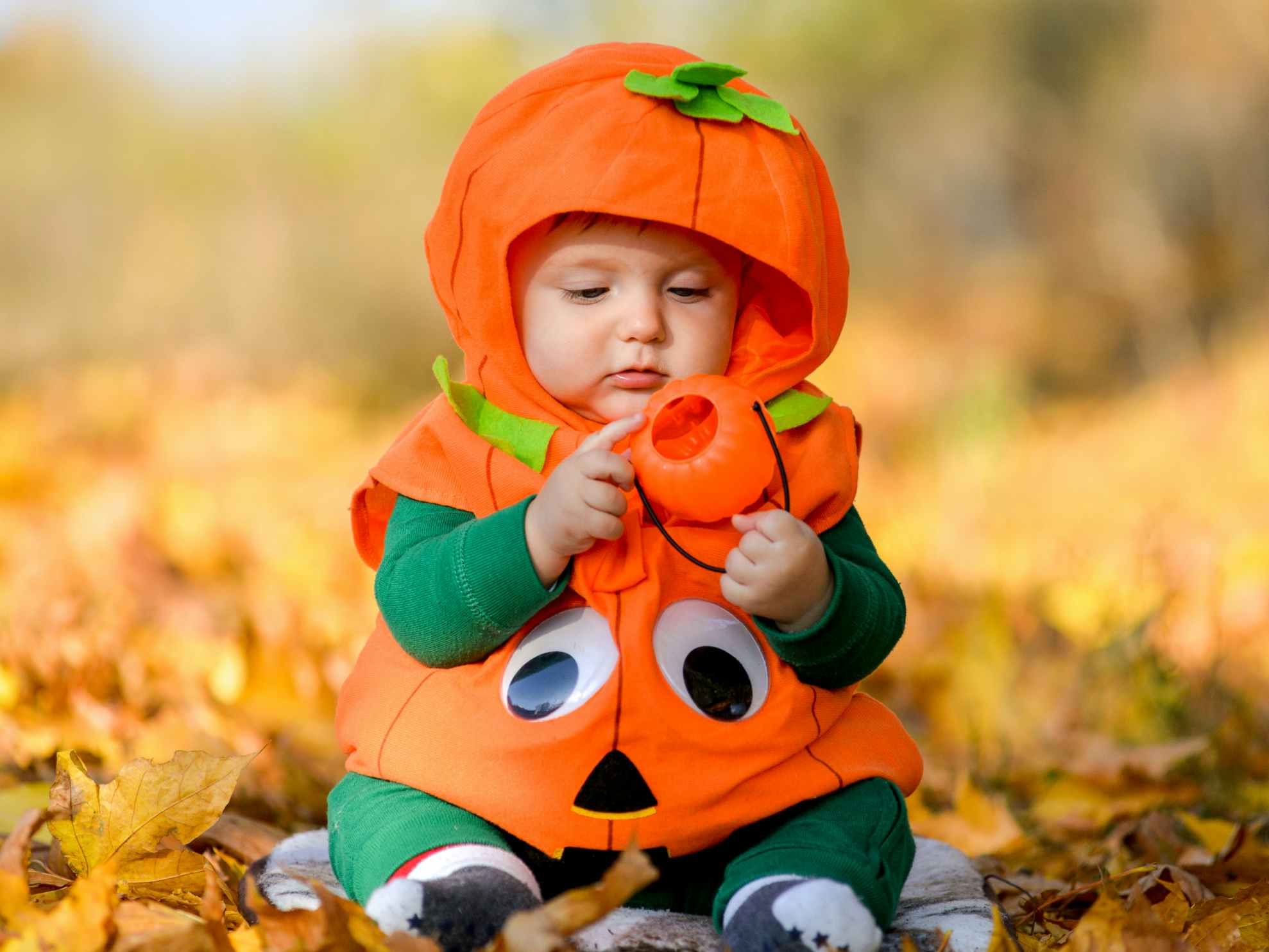 A baby dressed as a pumpkin sitting in some fallen leaves.
