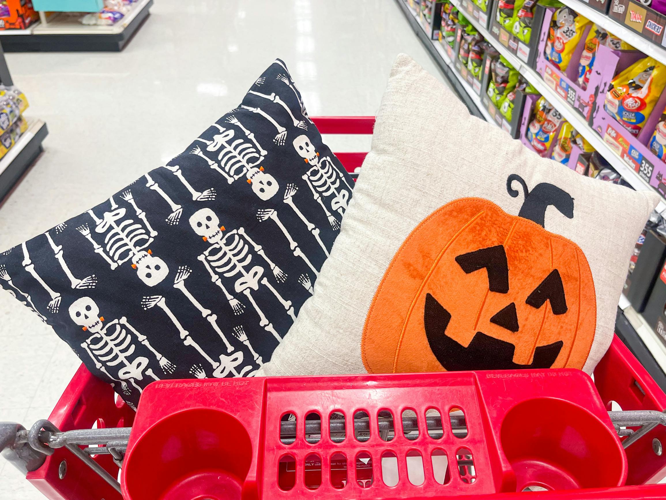 A throw pillow with skeletons and a throw pillow with a pumpkin on it are sitting in a Target shopping cart.