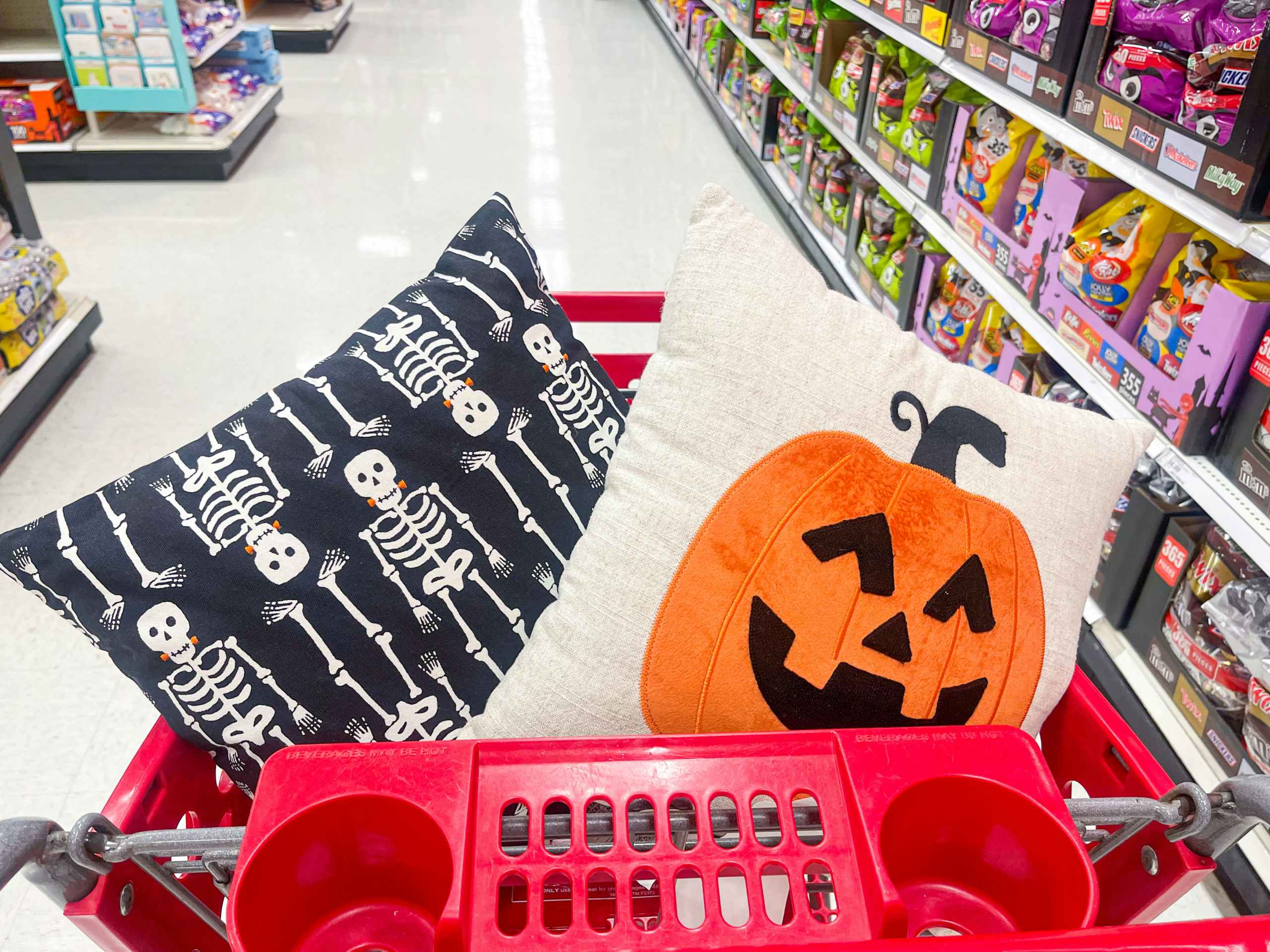 A throw pillow with skeletons and a throw pillow with a pumpkin on it are sitting in a store shopping cart.