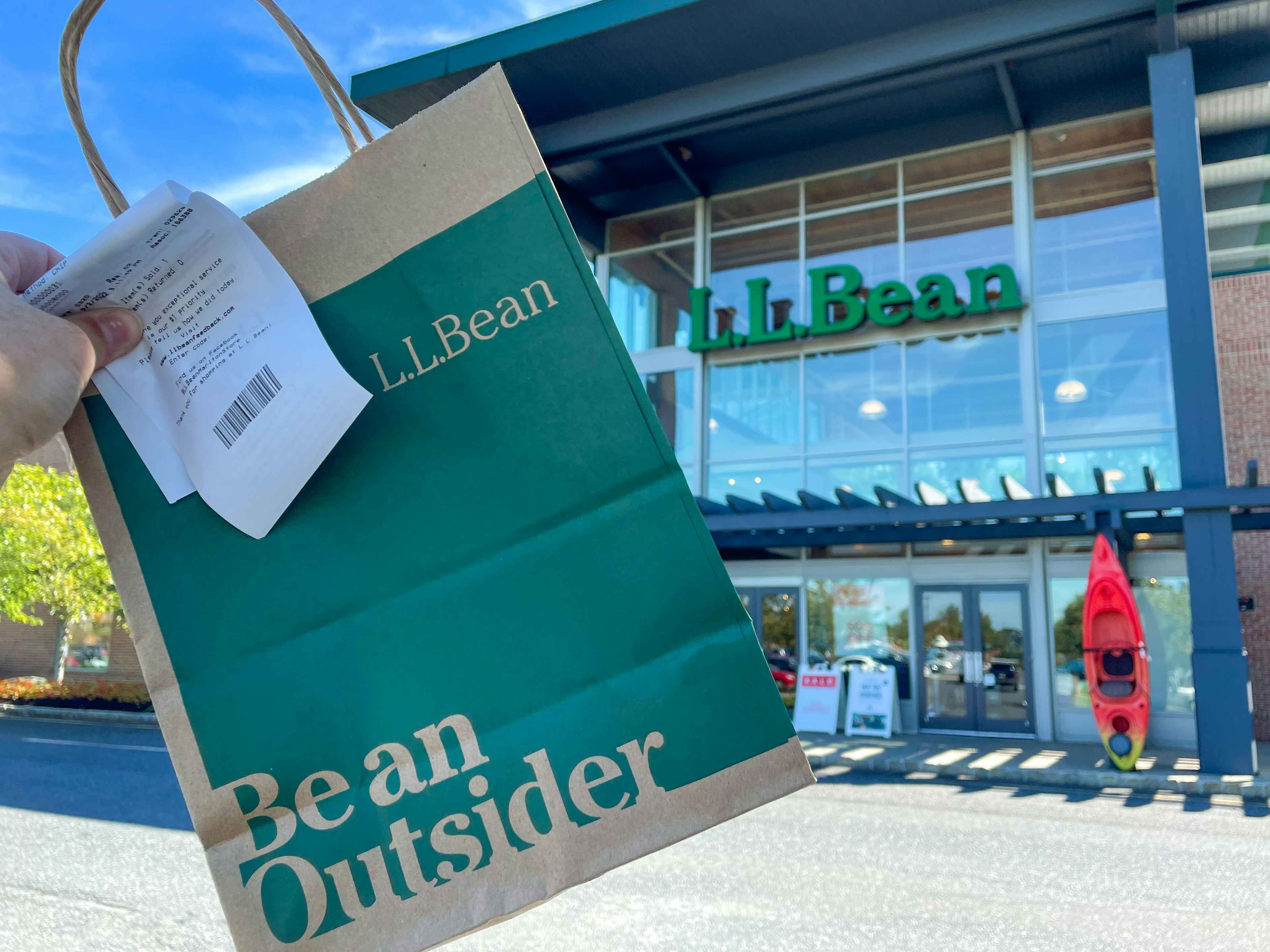 A person's hand holding up a L.L. Bean bag and receipt outside of an L.L.Bean store.