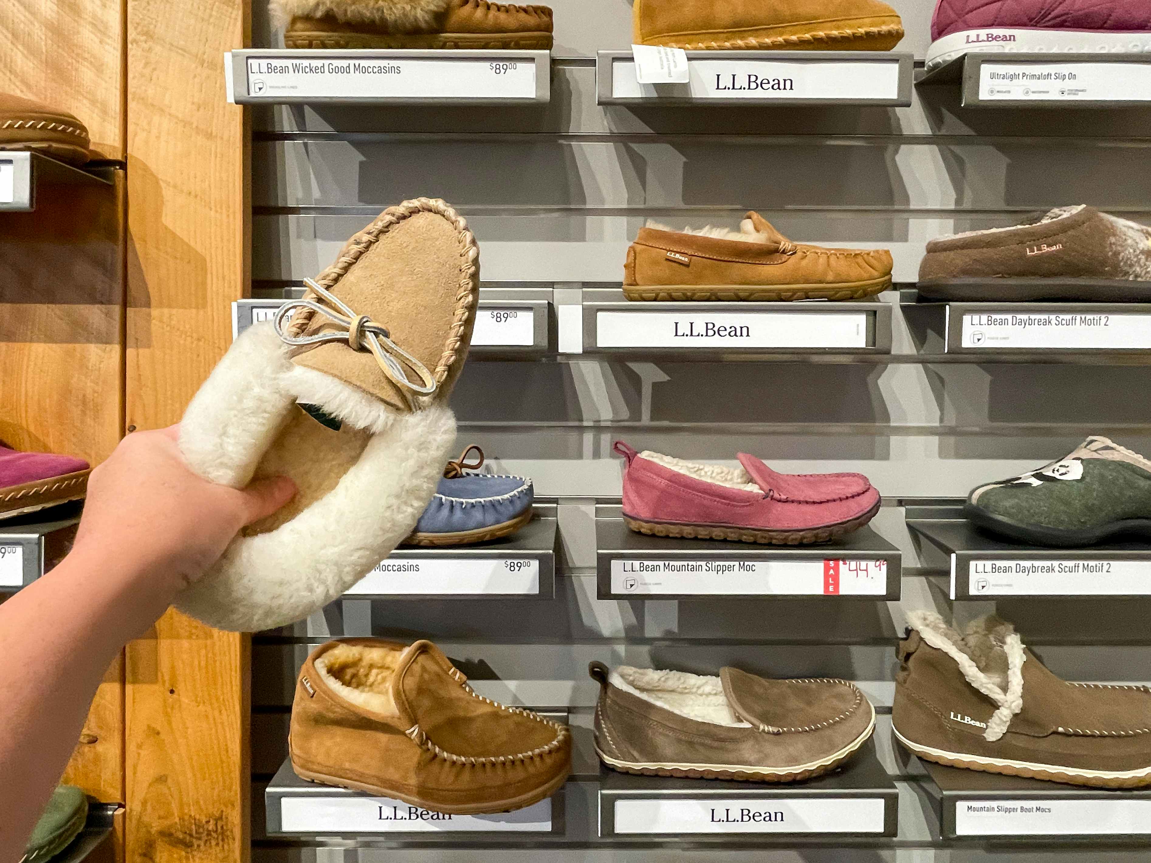 A person's hand holding up a Wicked Good Slipper in front of a wall of display moccasin slippers inside L.L.Bean