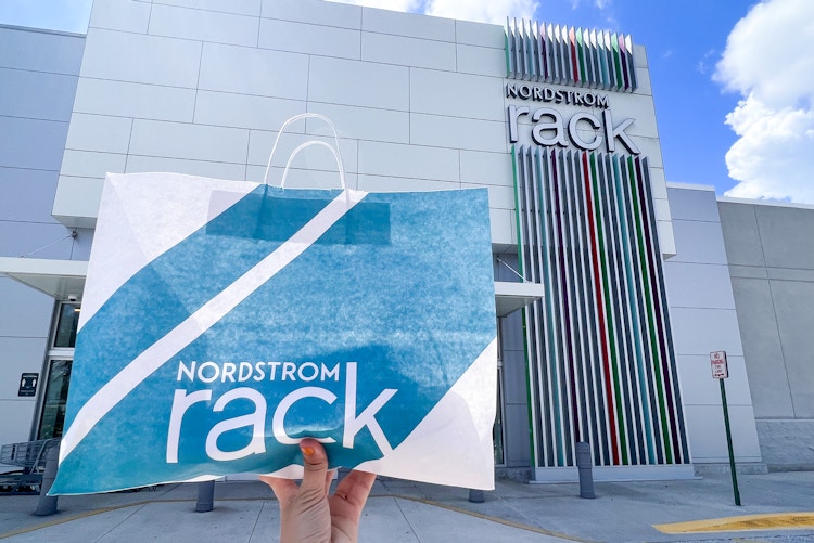 A person's hand holding up a Nordstrom Rack bag in front of a Nordstrom Rack store front.