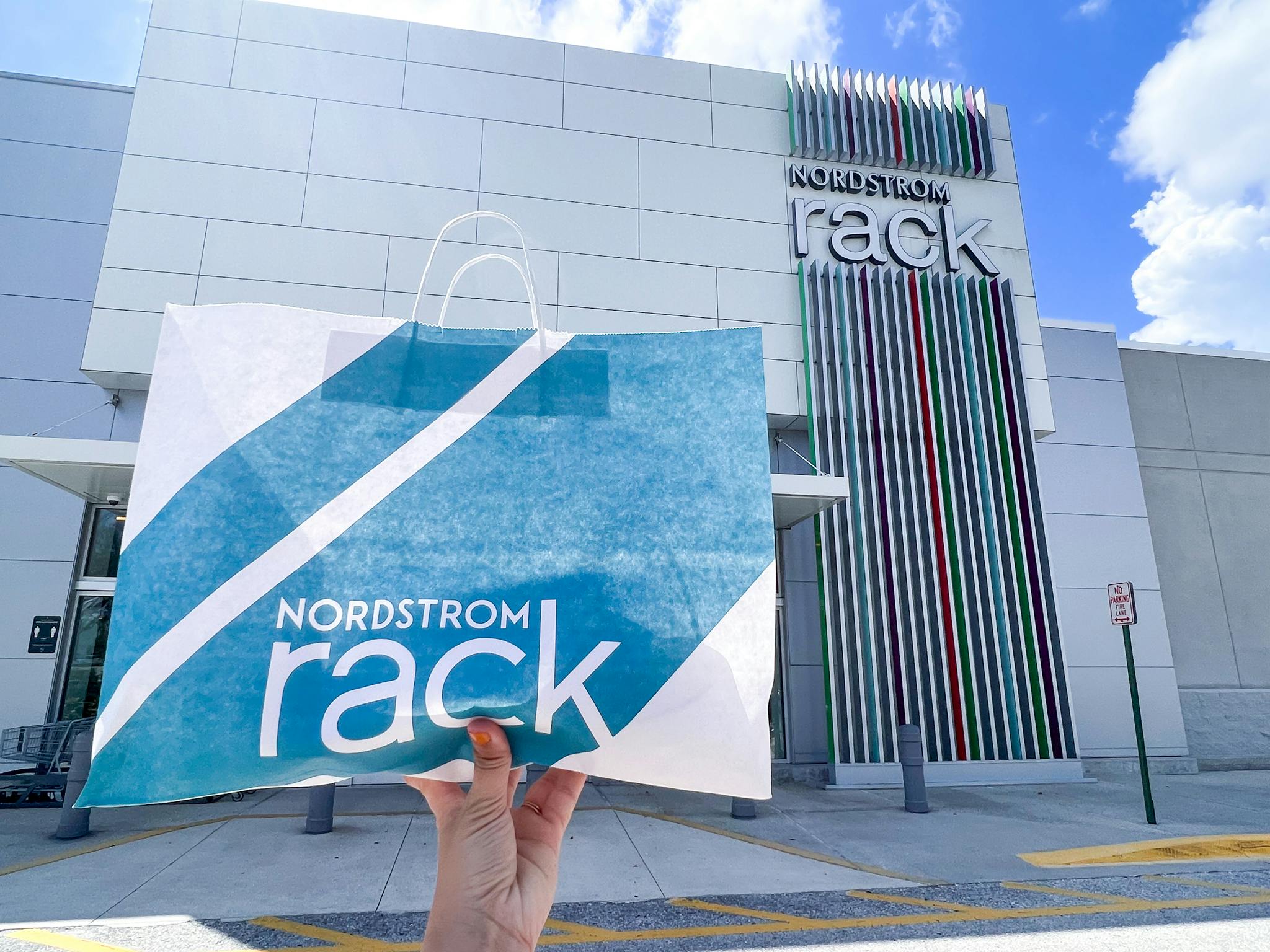 A person's hand holding up a Nordstrom Rack bag in front of a Nordstrom Rack store front.