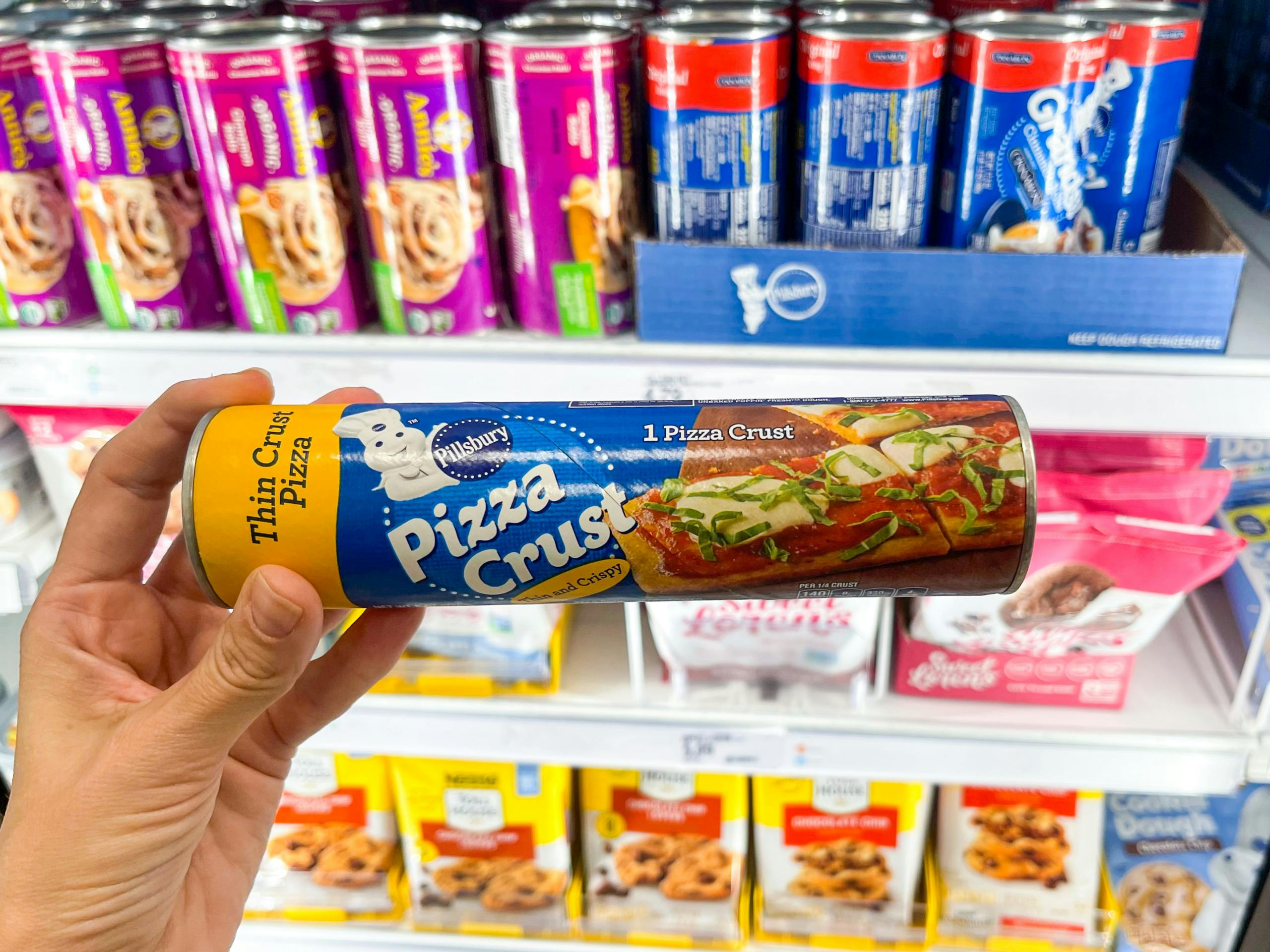 A can of pillsbury Thin crust pizza crust held out by hand in front of other food items sitting on the shelves in a freezer.