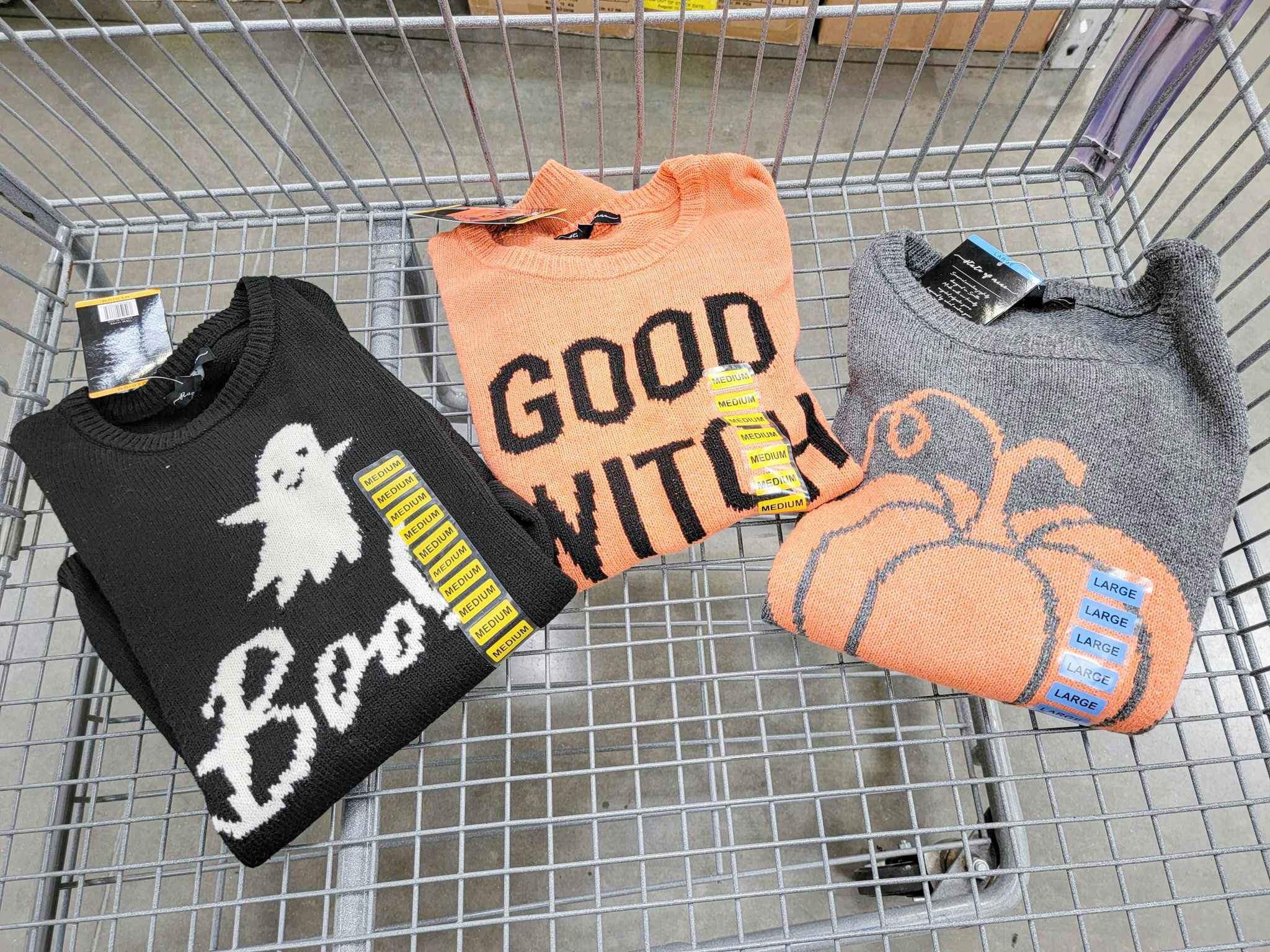 3 ladies halloween sweaters in a cart. Left one is black and says boo with a ghost on it, middle is orange and says good witch, right is grey with an orange pumpkin