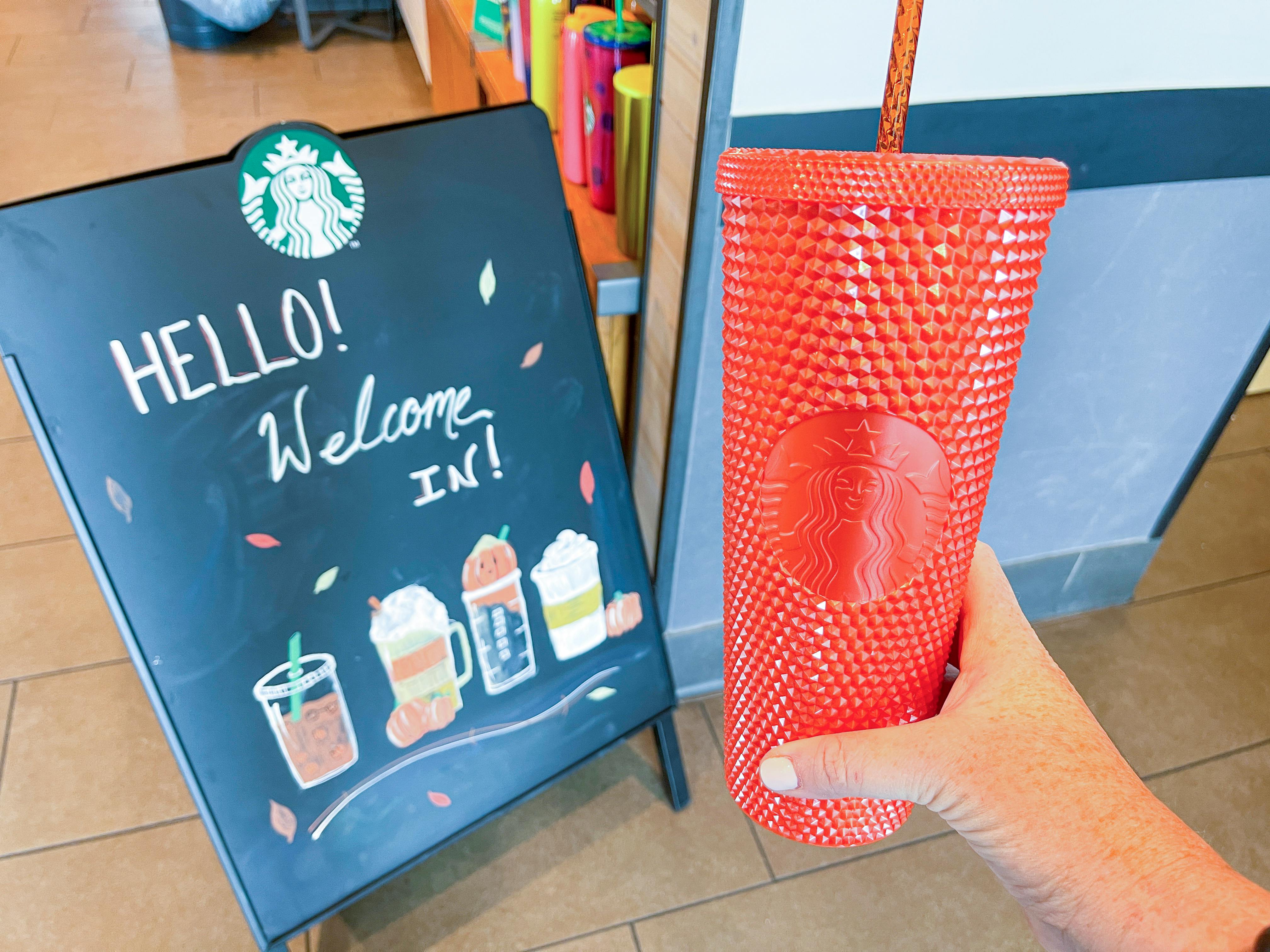 A person holding up a reusable Starbucks cup near a "welcome in" sign.