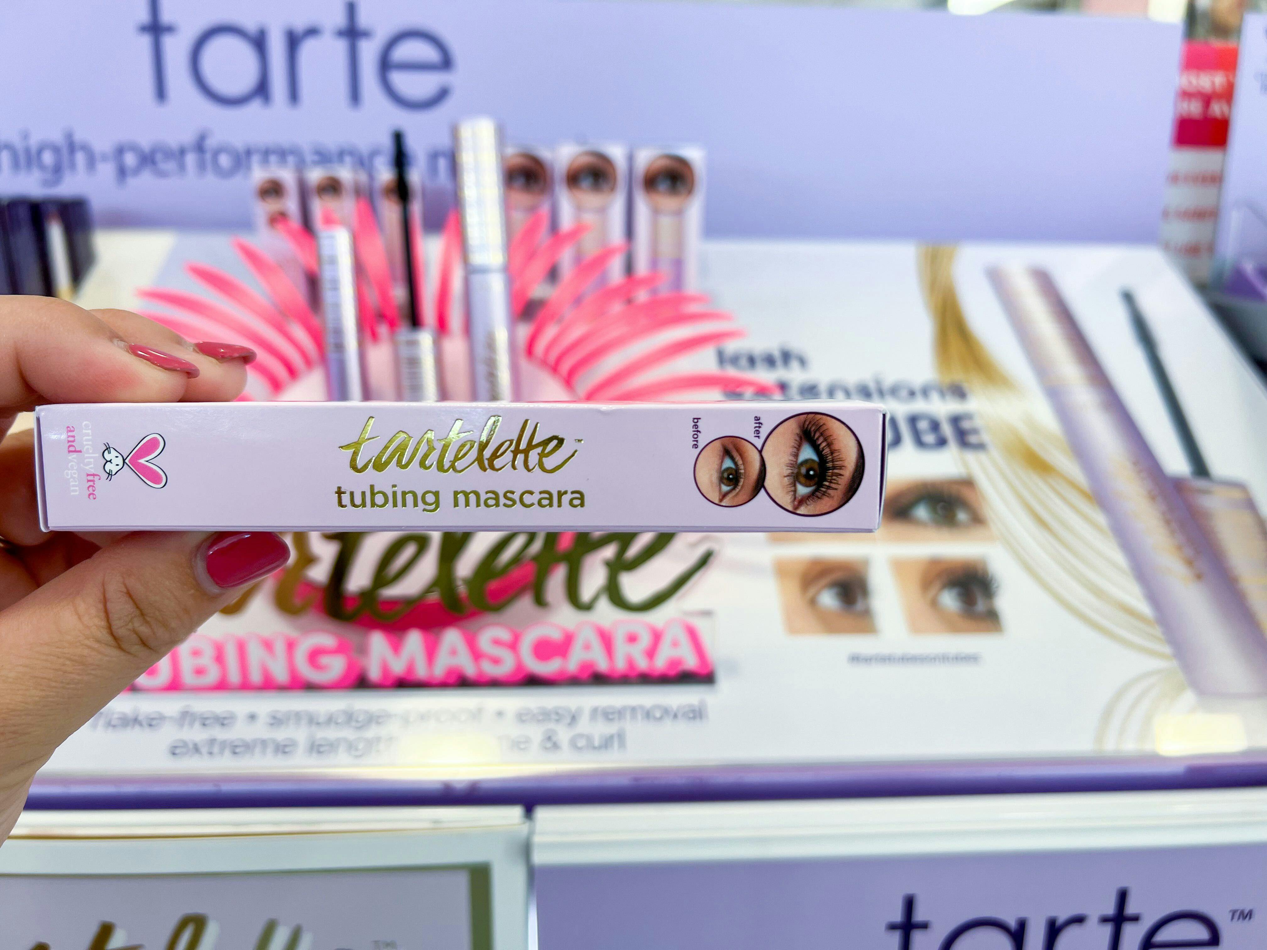 A hand holding a box of Tartelette tubing mascara in front of a pink and purple Tarte display