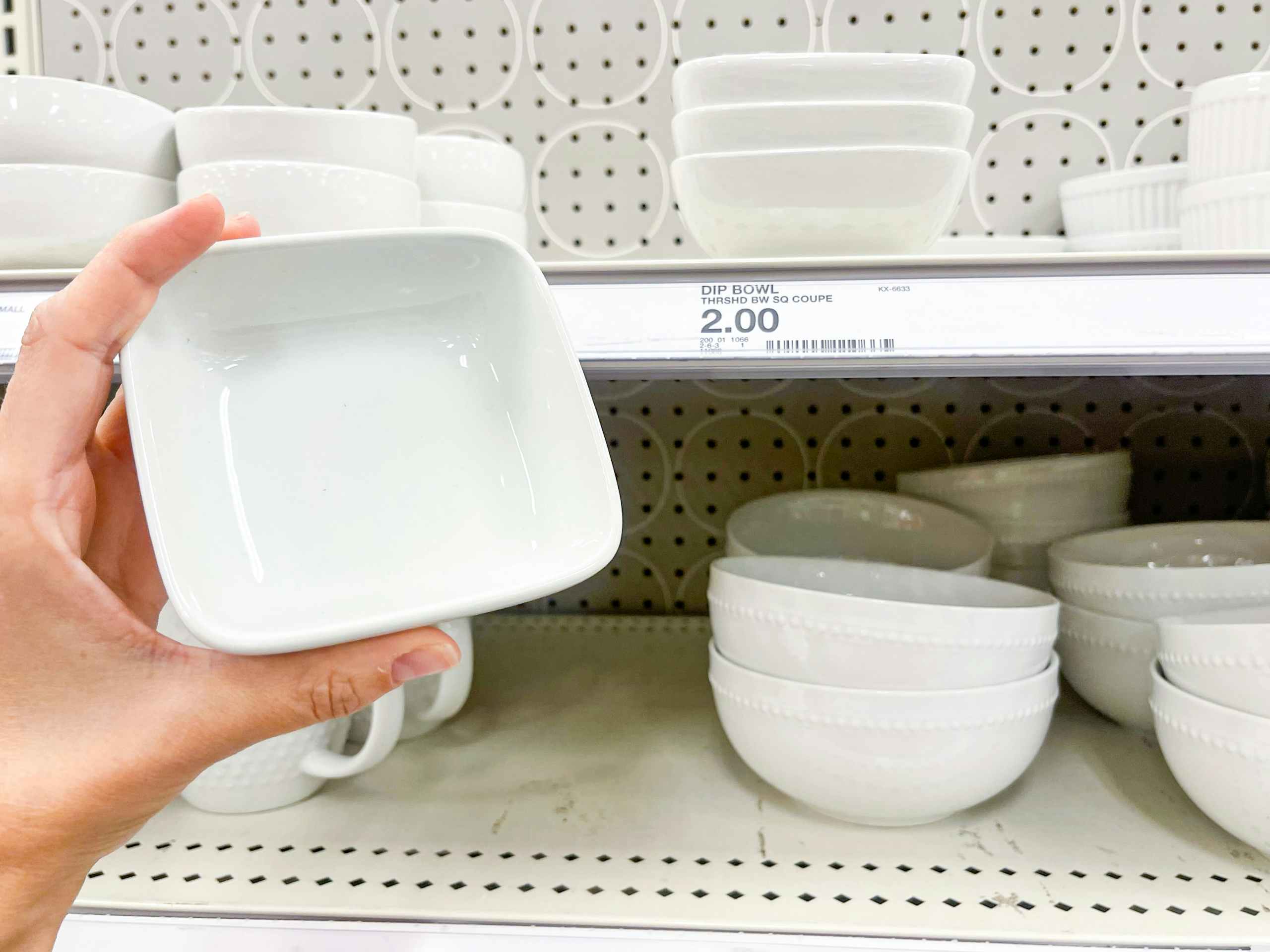 A small ceramic dip bowl held up with hand in front of price tag and other bowls sitting on a store shelf.