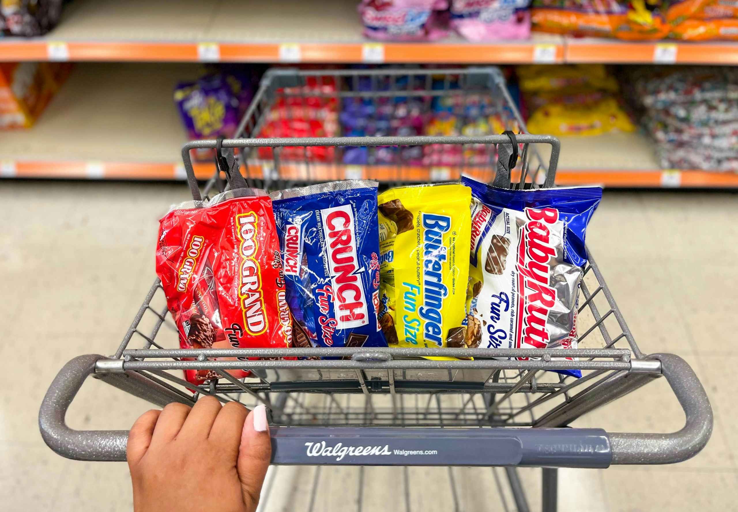 hand on shopping cart with a bag of Baby Ruth, Crunch, 100 Grand, and Butterfinger inside