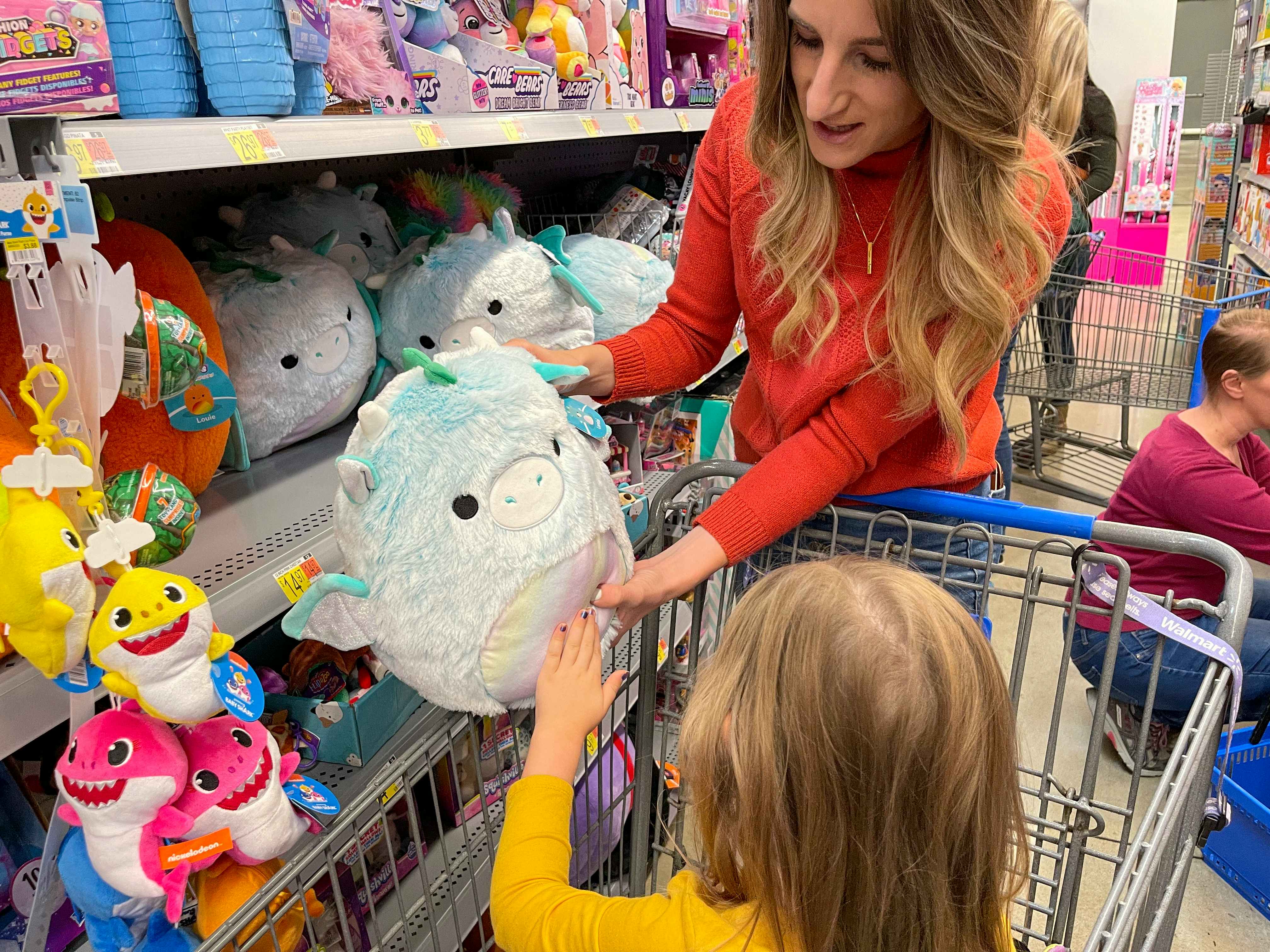 A woman showing a squishmallow to a little girl in a shopping cart.