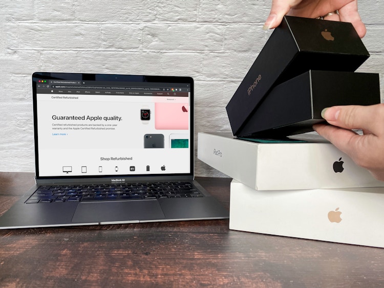 A person opening an old iPhone box next to a laptop displaying the Apple Refurbished products page.