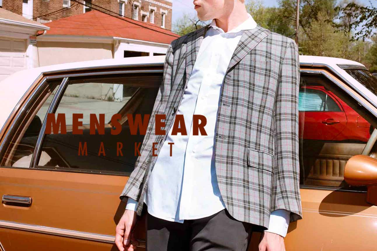 A man wearing a blazer leaning against an old car with the Menswear Market logo.
