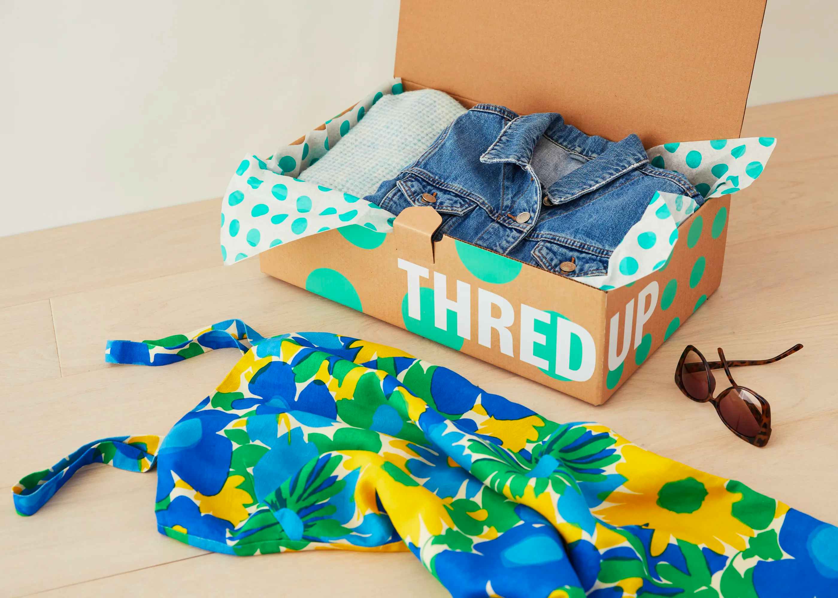 A ThredUp box with a denim jacket inside and some sunglasses and a dress on the table beside the box.