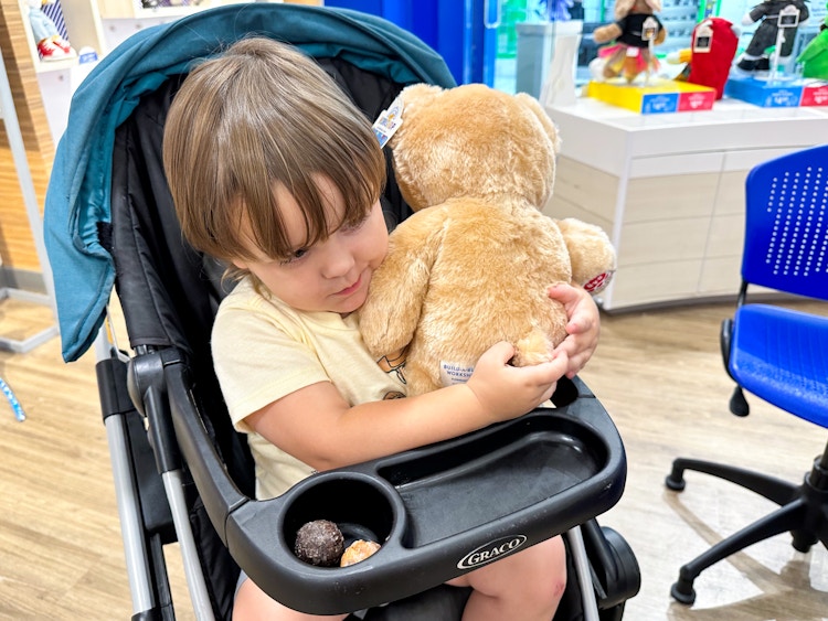 A little kid holding a Build-A-Bear Birthday Bear while sitting in a stroller