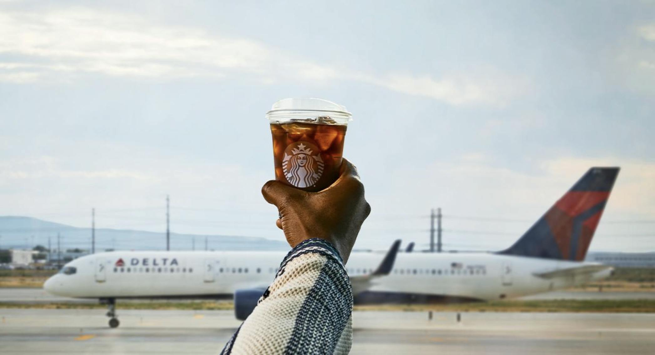 A hand holding a Starbucks coffee cup in front of a Delta Airlines plane